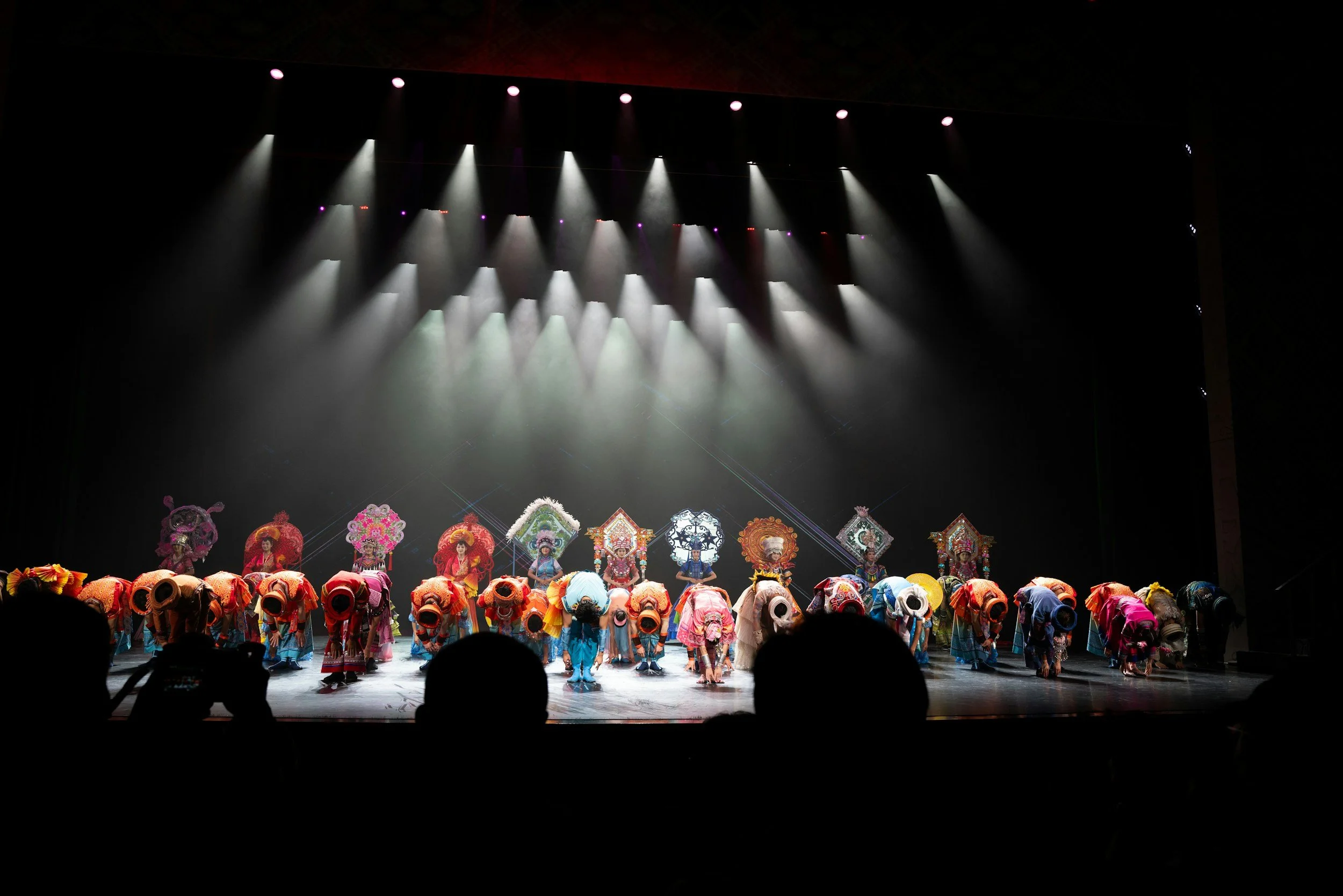 Stage performance with performers bowing, wearing colorful costumes and large ornate headpieces, under bright overhead stage lights.
