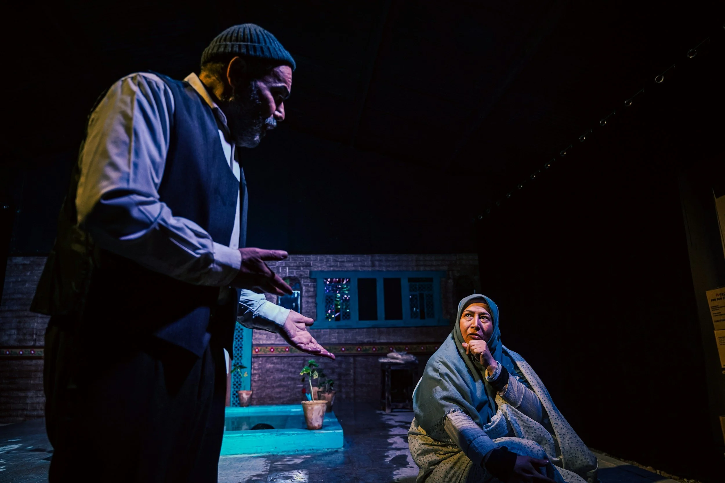 A man and woman are engaged in a theatrical performance on stage. The man, wearing a gray jacket and beanie, is standing and gesturing with his hands. The woman, dressed in traditional attire with a headscarf, is sitting on the stage and appears to be listening thoughtfully.