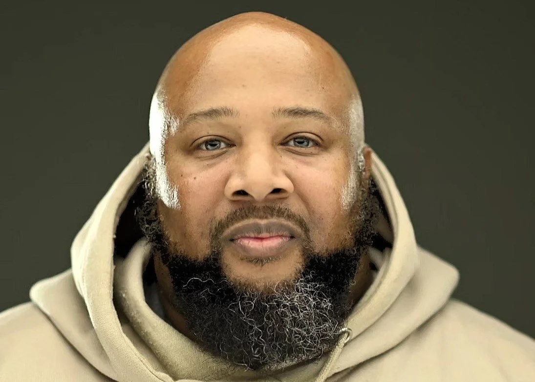 Close-up portrait of a bald man with a full beard wearing a beige hoodie against a dark background.