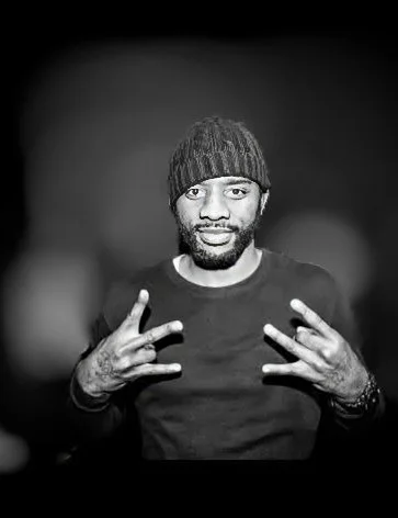 Black man wearing a knit beanie and a dark shirt, making peace signs with both hands, in a black-and-white photo.