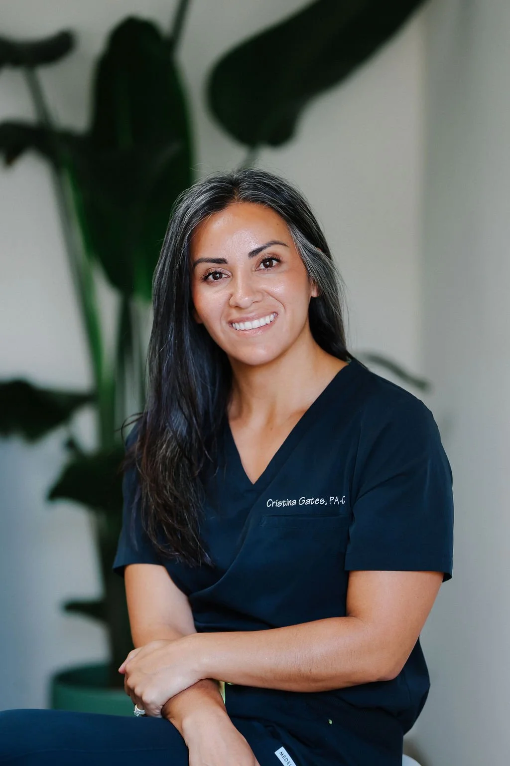 A woman with long dark hair and a warm smile, wearing navy scrubs with embroidered name "Cristina Gates, PA-C," sitting in front of a plant with large green leaves.
