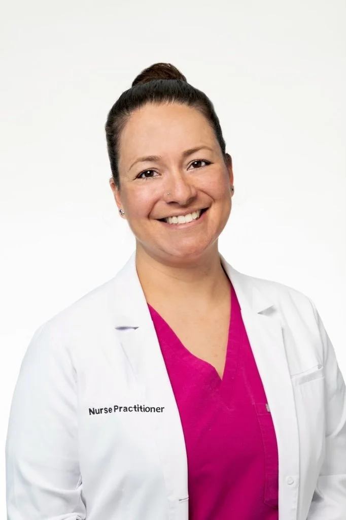 A smiling nurse practitioner in a white coat with pink scrubs against a white background.