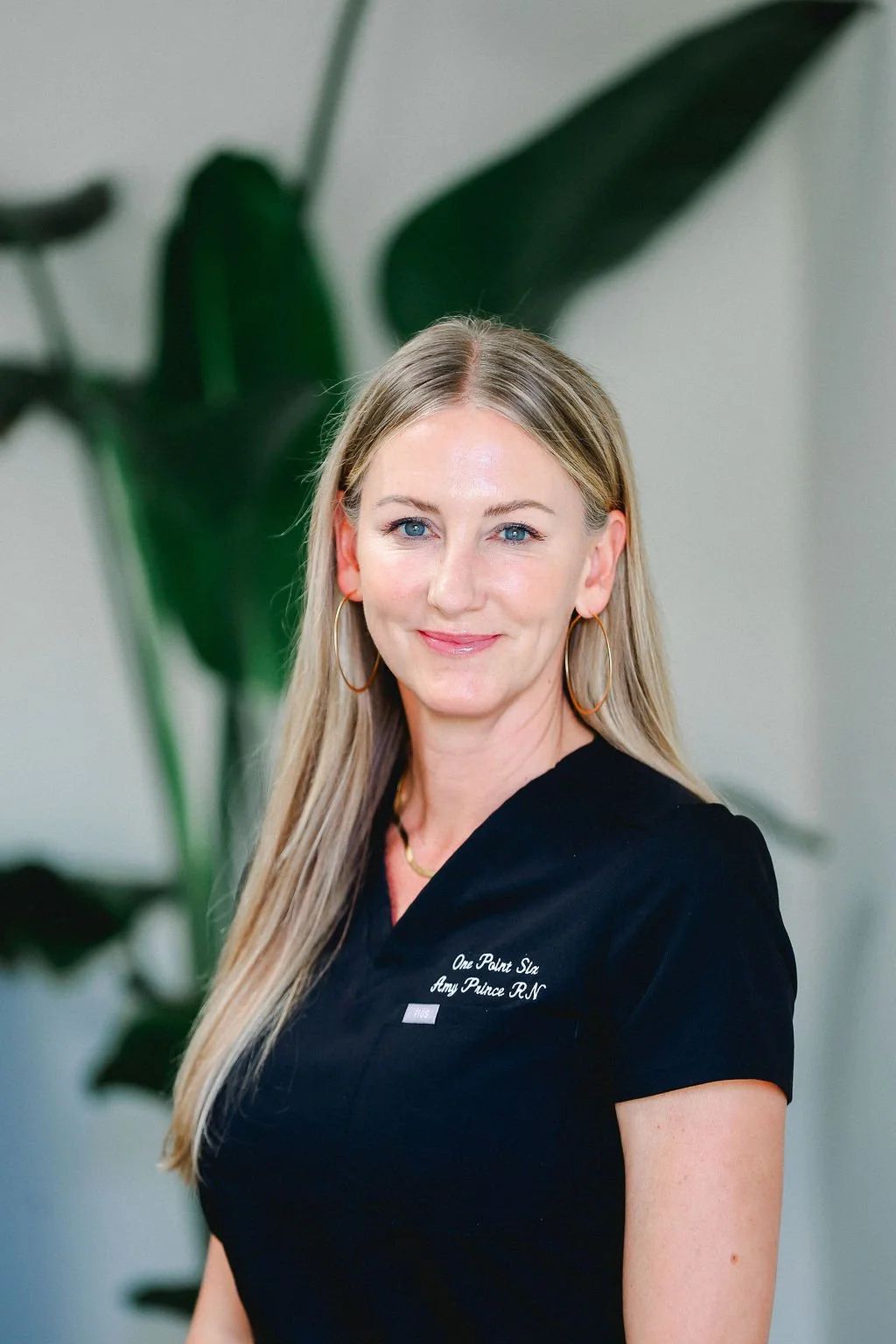 A smiling woman with long blonde hair, wearing a black medical uniform with 'One Point Spa Amy Prince R.N.' embroidered on it, standing in front of a large green leafy plant.