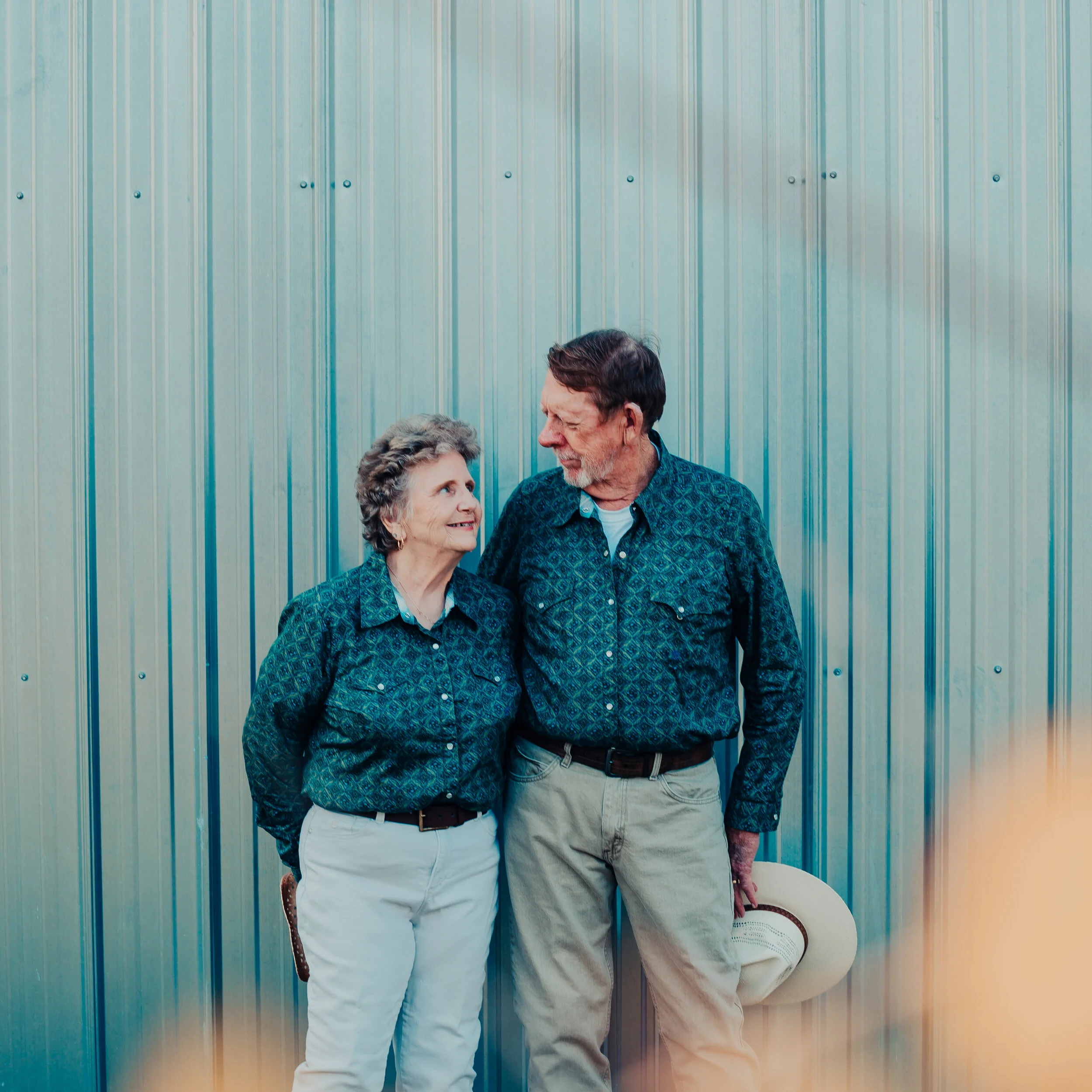 An elderly couple wearing matching patterned blue shirts standing close together in front of a metallic wall, smiling at each other, with the woman holding a brown hat.