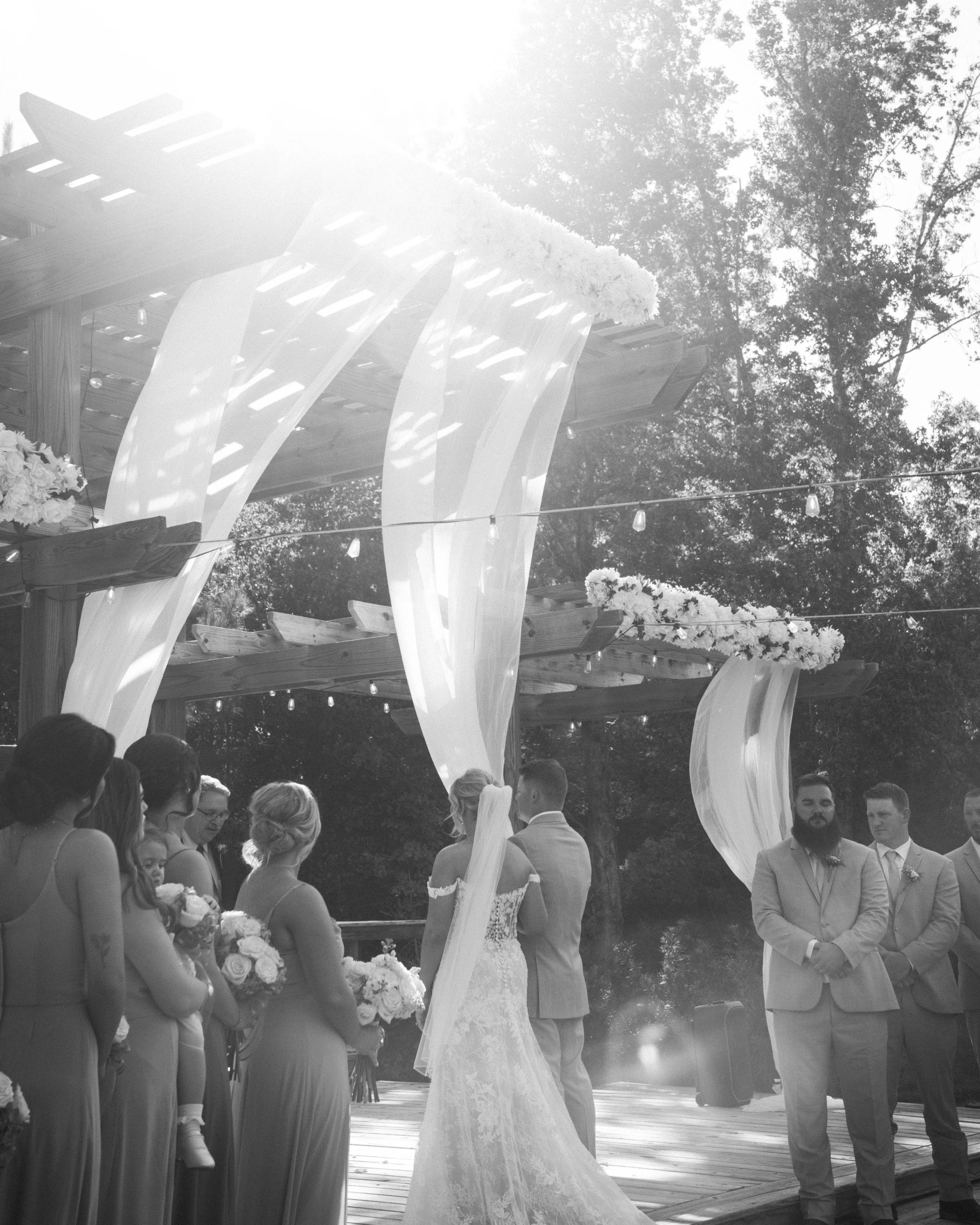 A black and white photo of a wedding ceremony on an outdoor stage with flowing white fabric and floral decorations, with the bride and groom standing together and guests watching.