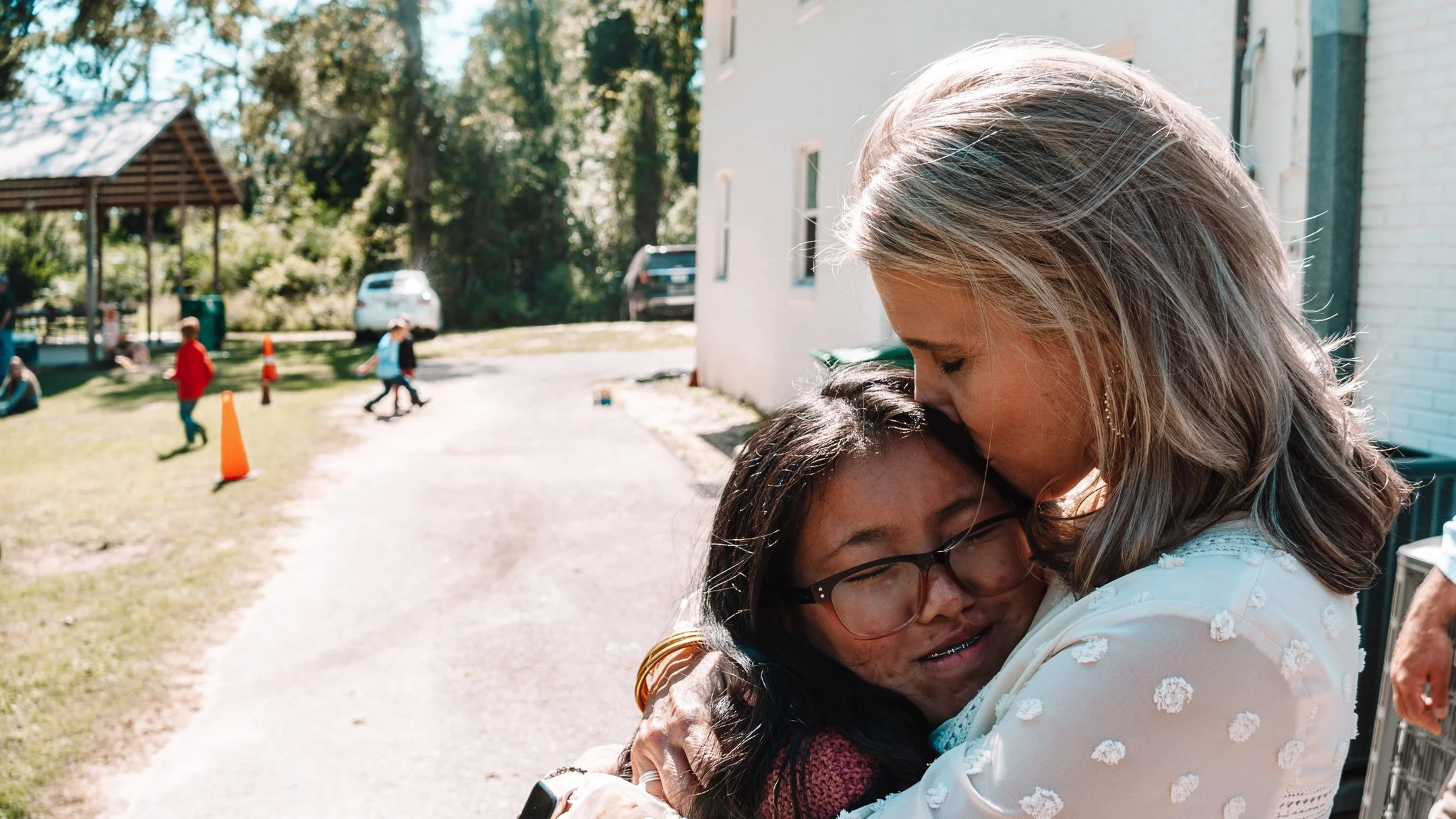 A woman is hugging a girl with glasses outdoors on a sunny day, with children playing in the background near orange cones and a pavilion.