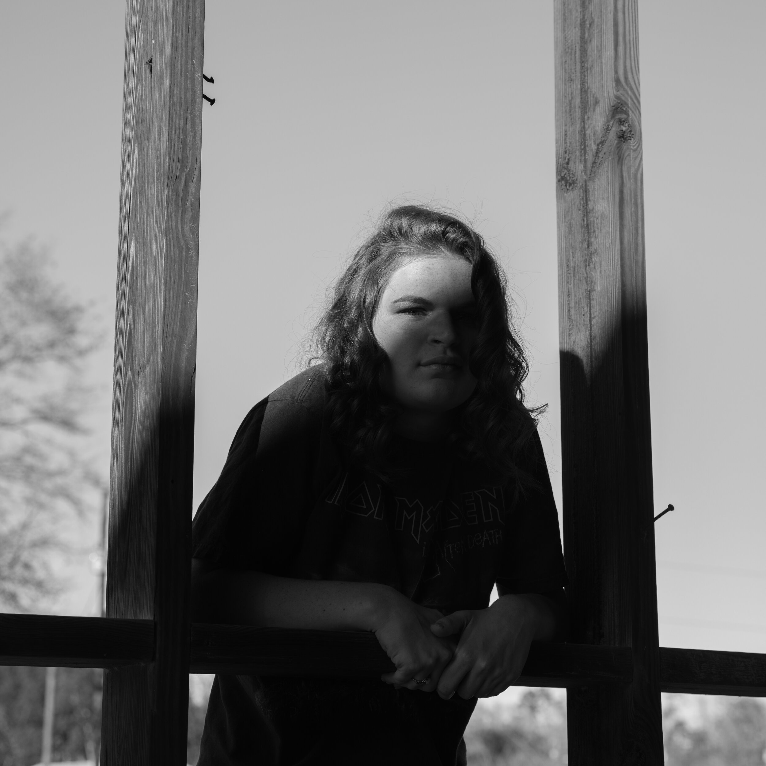 Black and white photo of a young woman with wavy hair leaning on a wooden structure outdoors, with a blurred background of trees and sky.