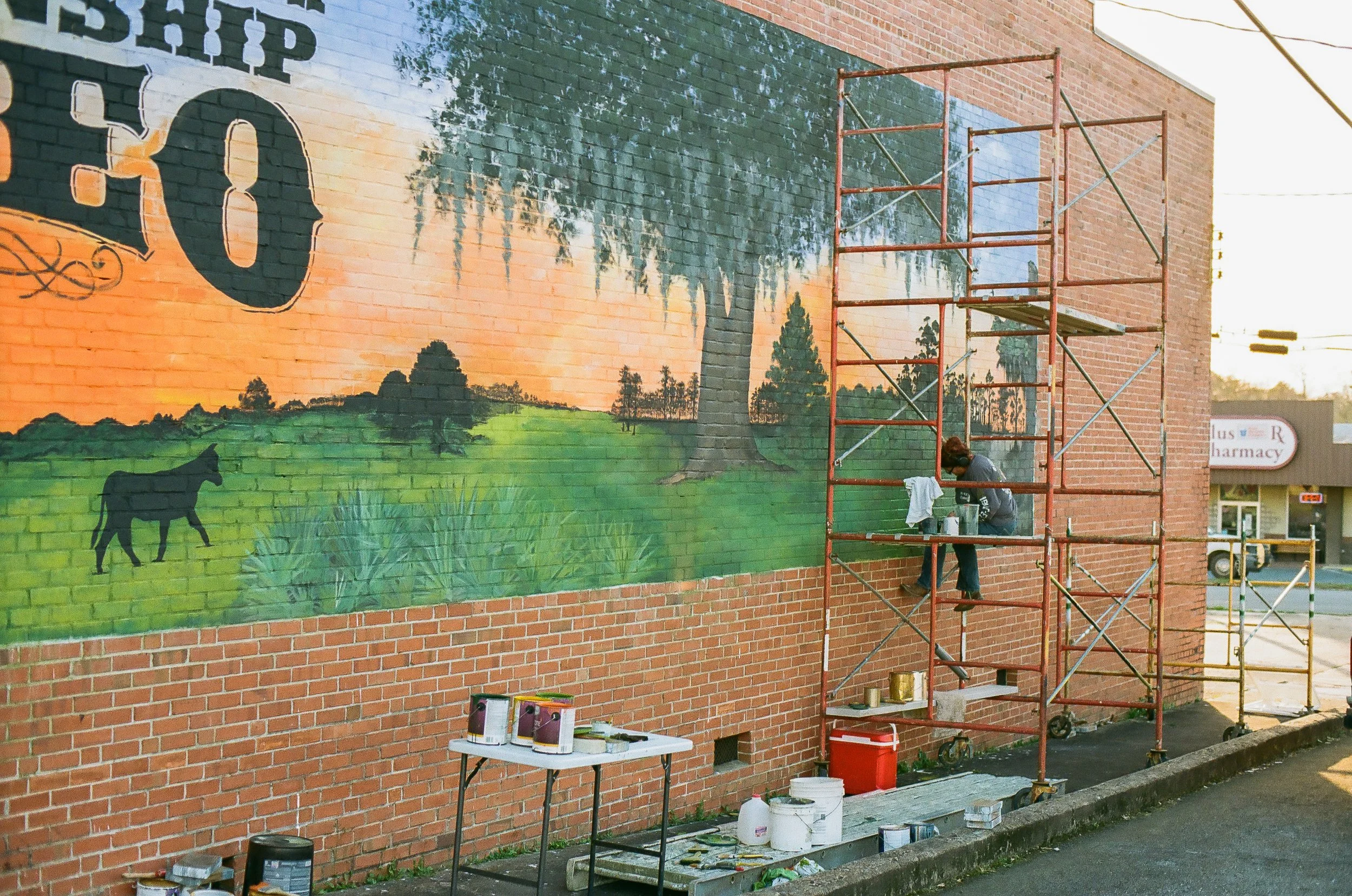 A person painting a mural on a brick wall using scaffolding, with a table and paint supplies on the ground.