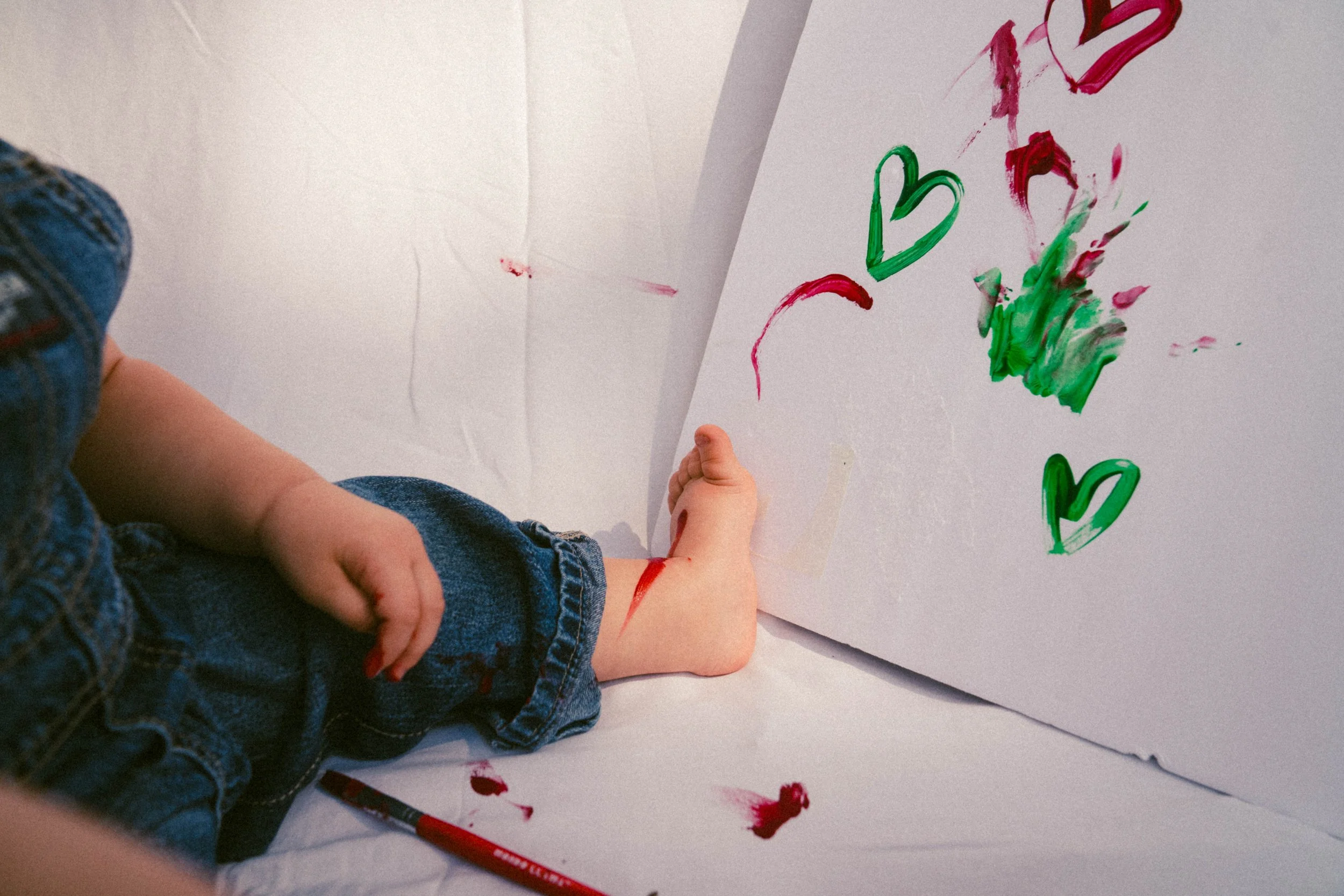 Child's hand and legs with painted red and green hearts on white paper background, some paint smears, and a red paintbrush on the floor.
