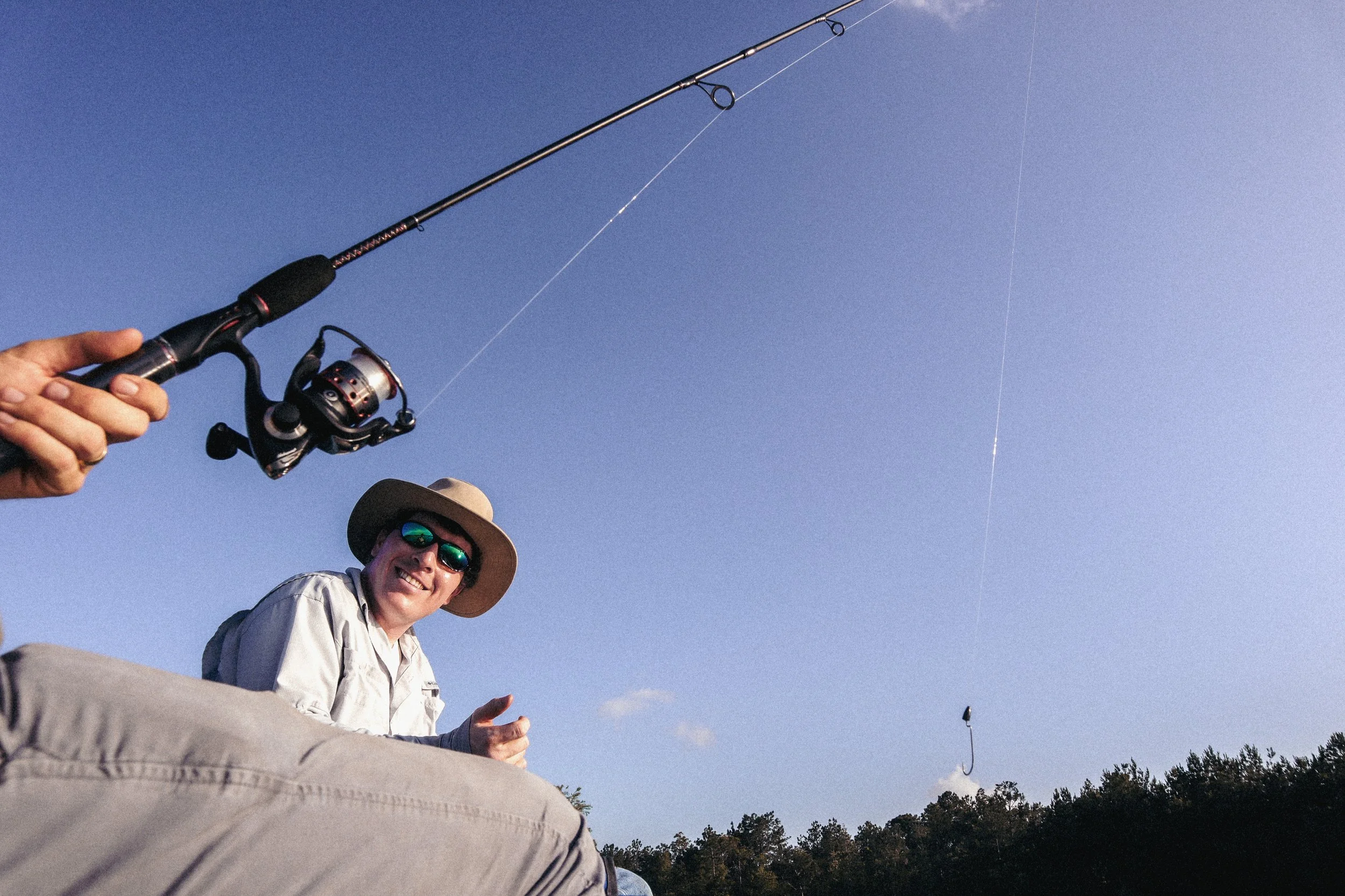 A man smiling and giving a thumbs-up while fishing with a spinning rod in an outdoor setting with a clear blue sky and trees in the background.