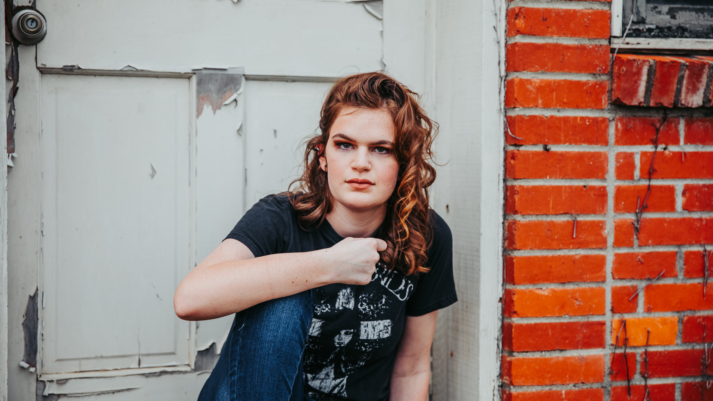 Young woman with curly red hair, wearing a black t-shirt and blue jeans, crouching outside next to a weathered white door and red brick wall.