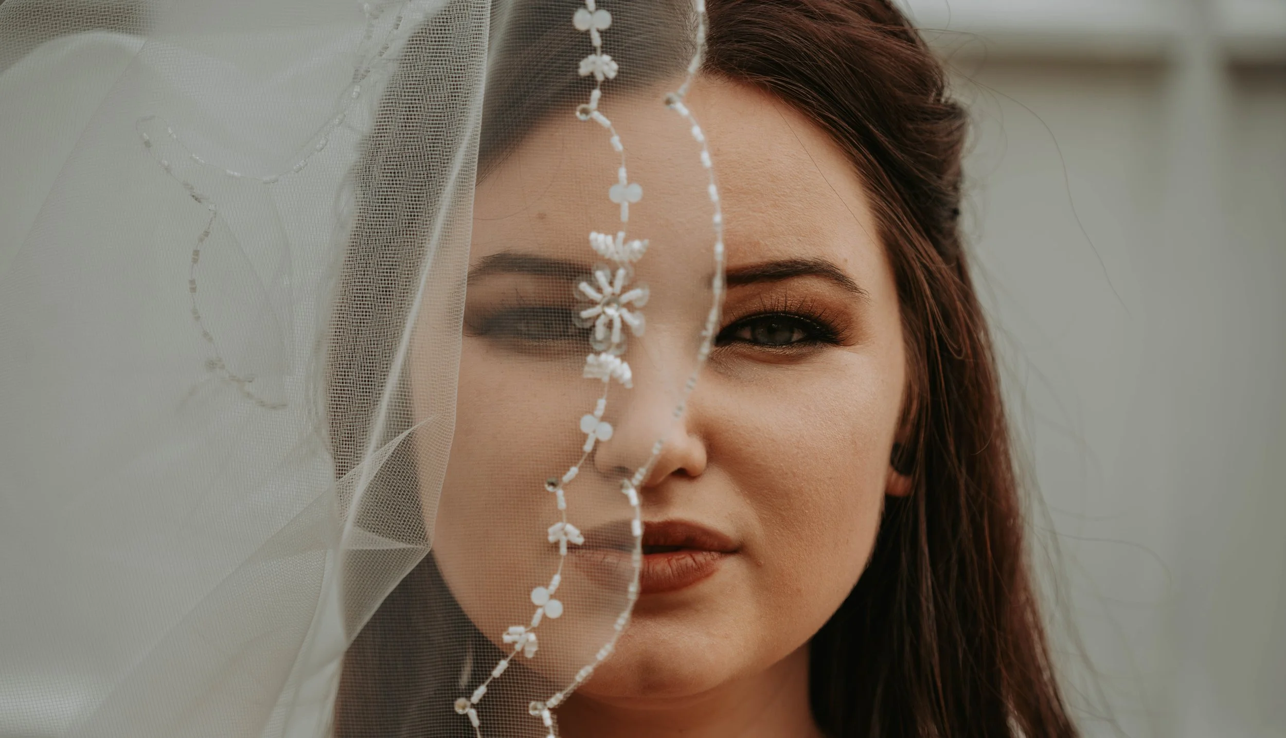 Close-up of a woman with long brown hair and makeup, partially covered by a sheer white veil with embroidered details, outdoors.