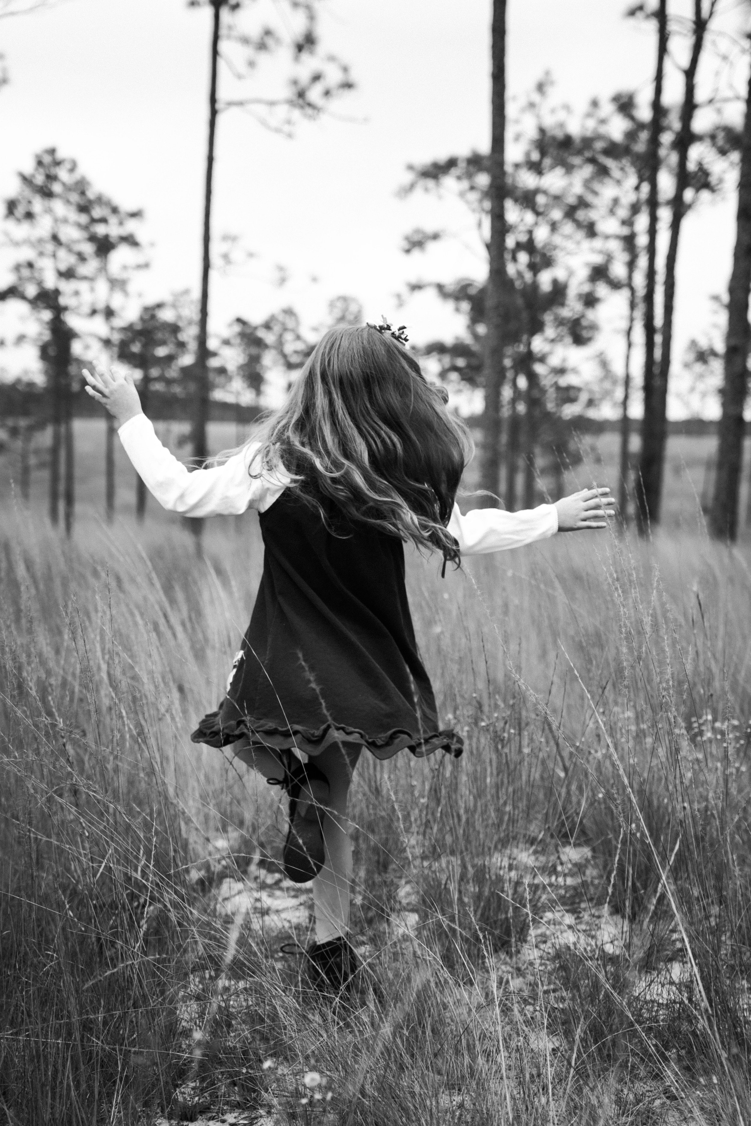 A girl with long hair wearing a black dress with ruffled hem, a white blouse, and dark shoes, running through tall grass in a wooded outdoor area on a cloudy day.