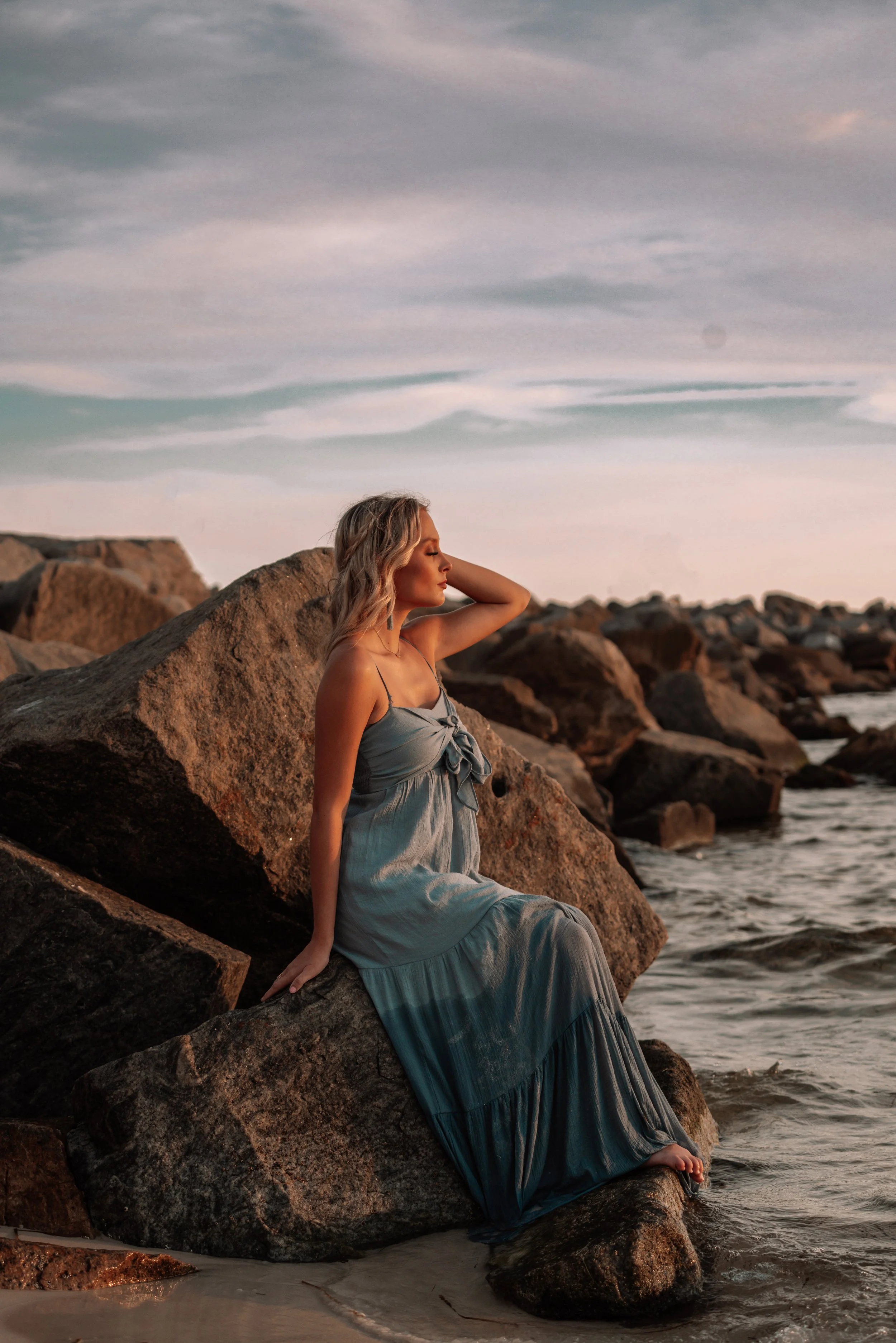 A woman in a blue dress sitting on a large rock by the shoreline during sunset.