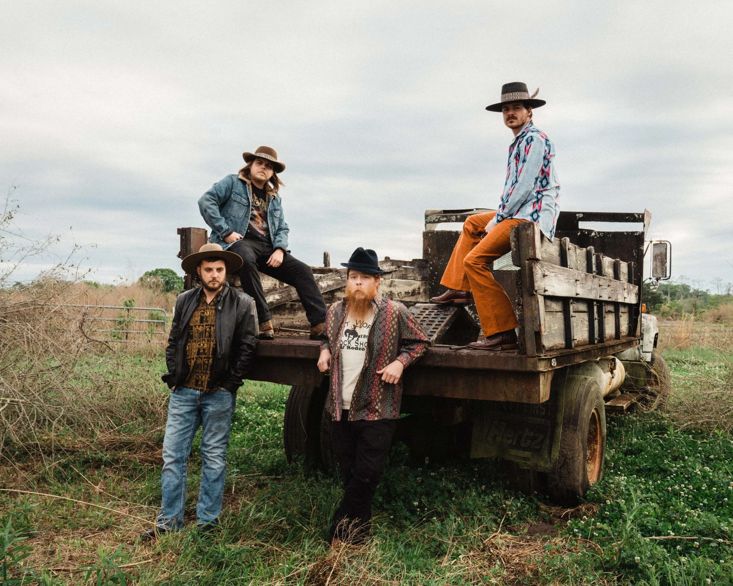 Five men with beards and hats posing on and around a weathered flatbed truck in an open field with overcast sky.