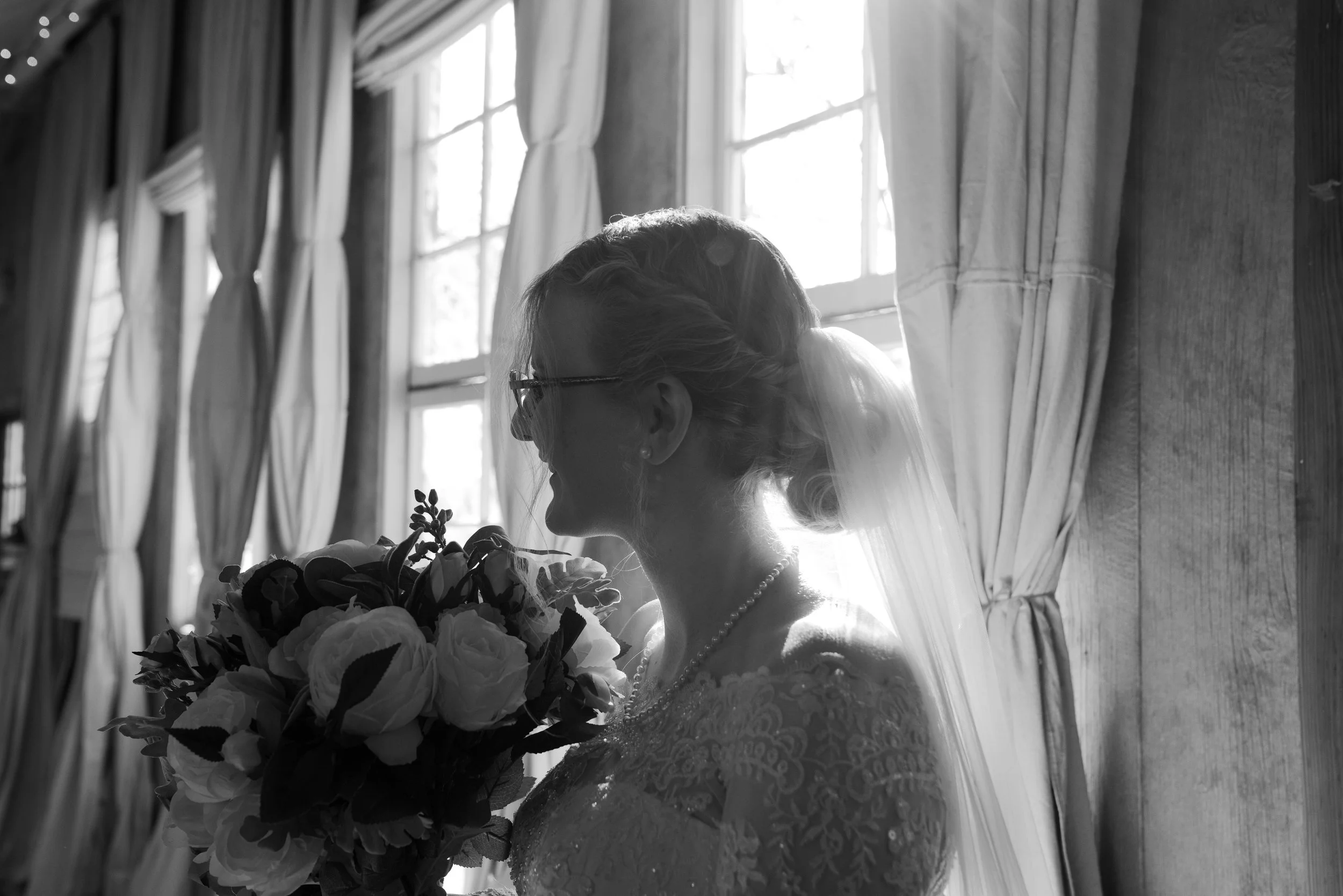 A bride holding a bouquet of flowers, wearing glasses and a lace wedding dress, standing indoors by a window with curtains, backlit with natural light.