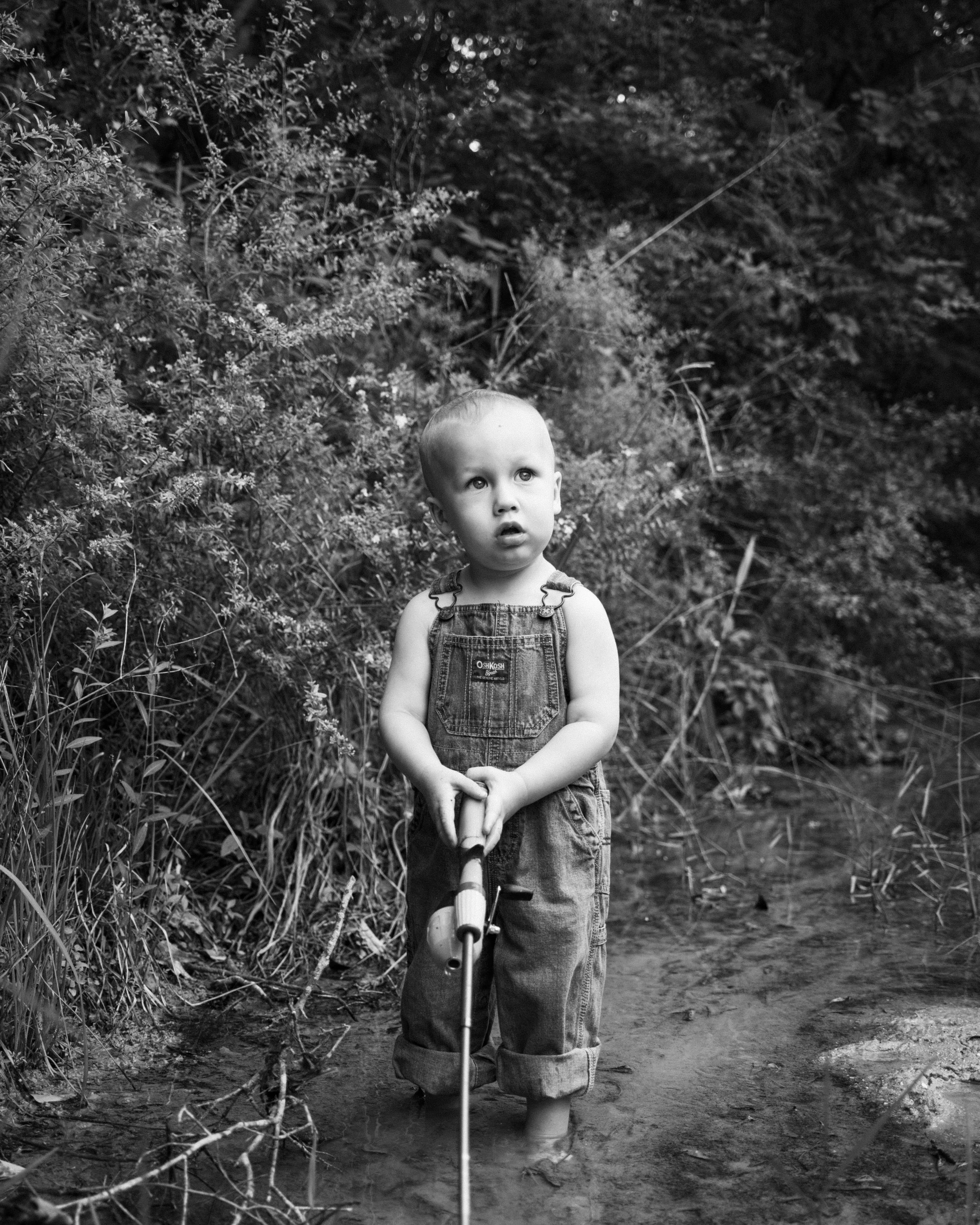 A young child standing in shallow water outdoors, holding a stick or toy, surrounded by dense foliage, in black and white.