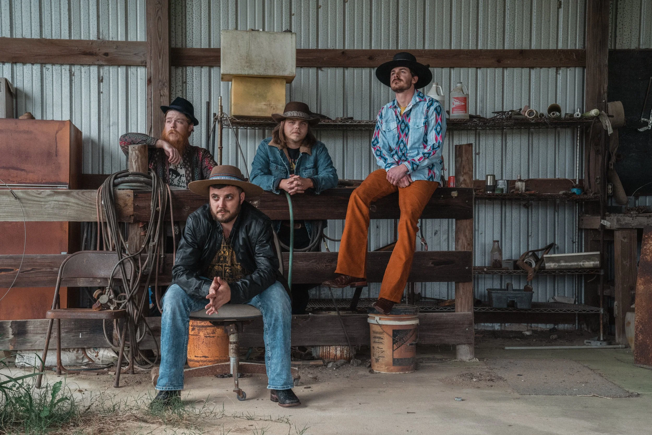 Four men sitting and standing in a rustic barn or workshop, wearing eclectic clothing including cowboy hats and colorful shirts, with tools and equipment around them.