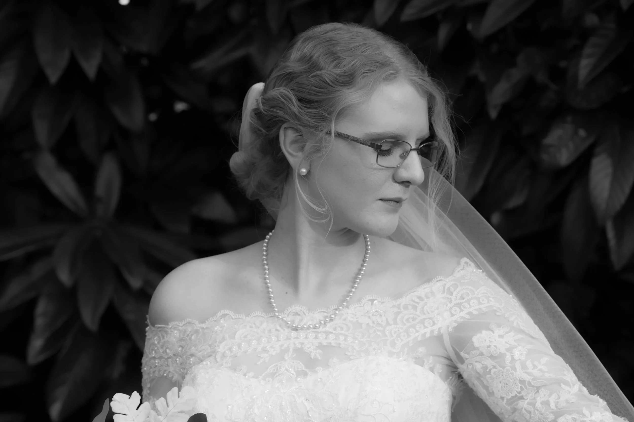 Black and white photo of a bride with glasses, wearing a lace wedding dress, pearl necklace, and veil, looking down while standing in front of leafy plants.