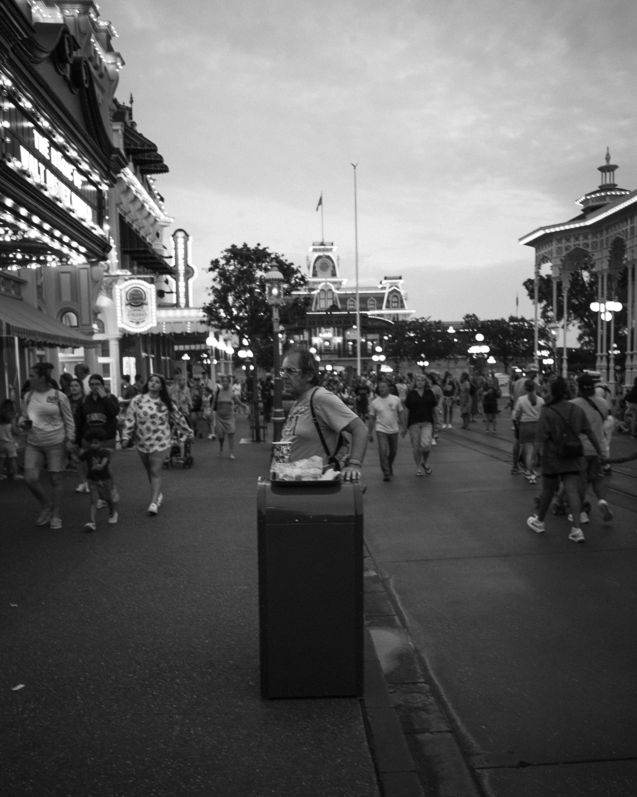 Black and white photo of a crowded amusement park entrance with people walking, some pulling luggage or carrying bags, and a man standing next to a trash can in the foreground.