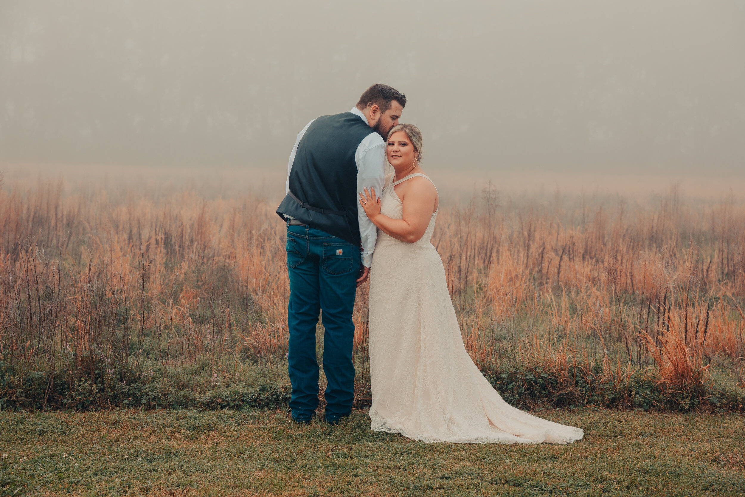 A couple dressed in wedding attire standing close together outdoors on a foggy day, with the woman in a white lace wedding gown and the man in a white shirt with a dark vest and jeans, holding hands and touching foreheads.