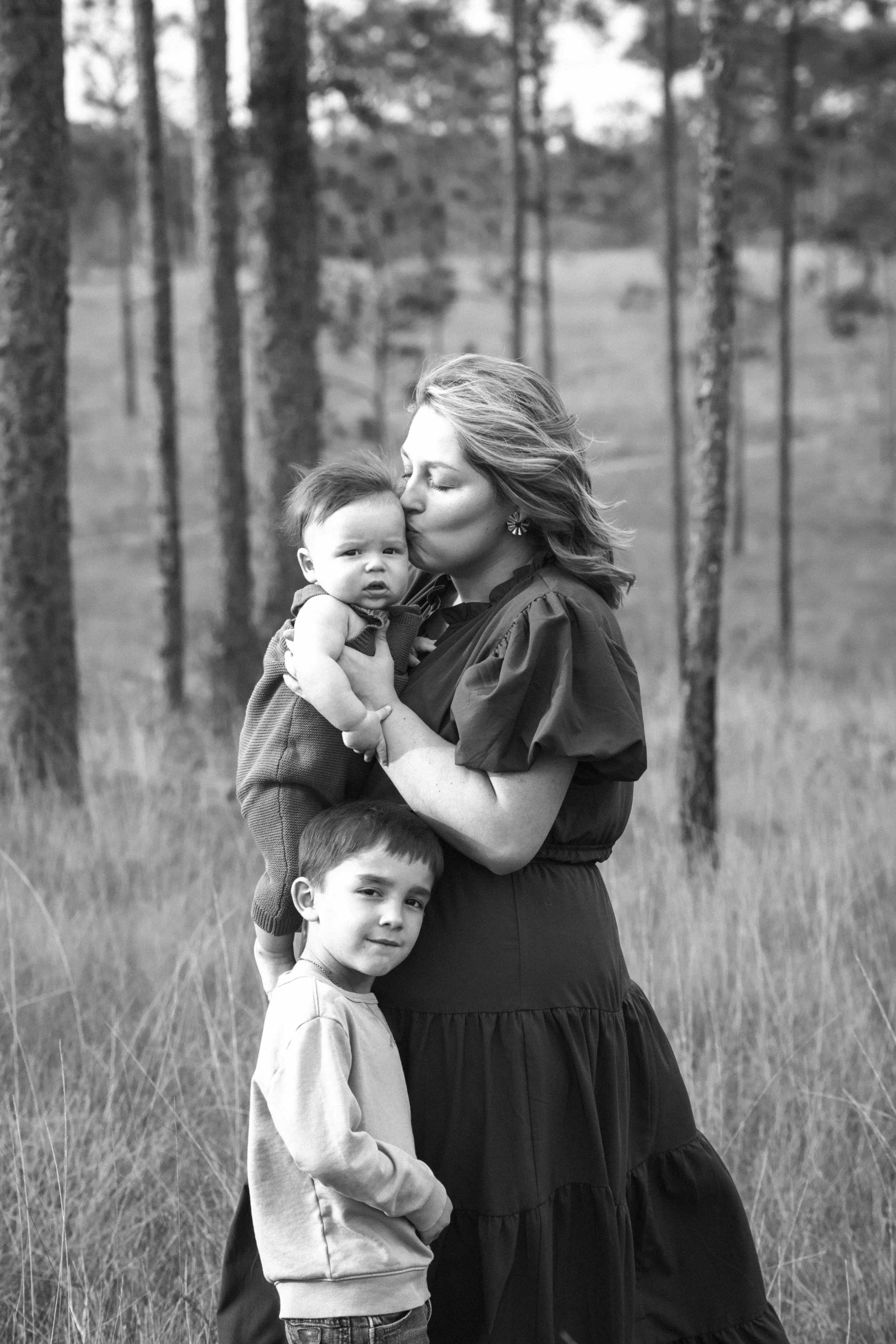 A woman holding a baby in a forest, with a young boy standing next to her, in black and white.