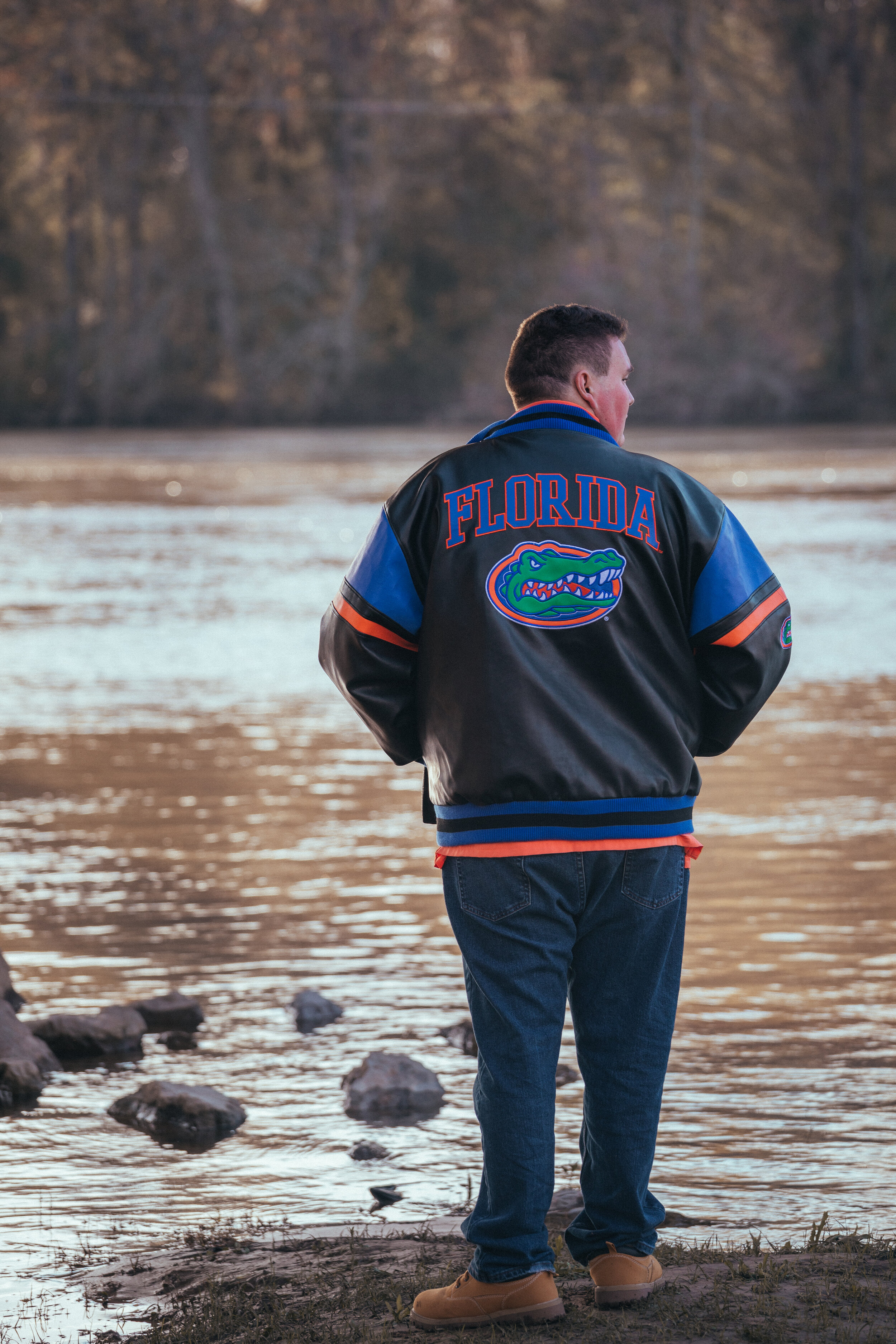 A man standing by a river, wearing a Florida Gators jacket, looking away from camera.