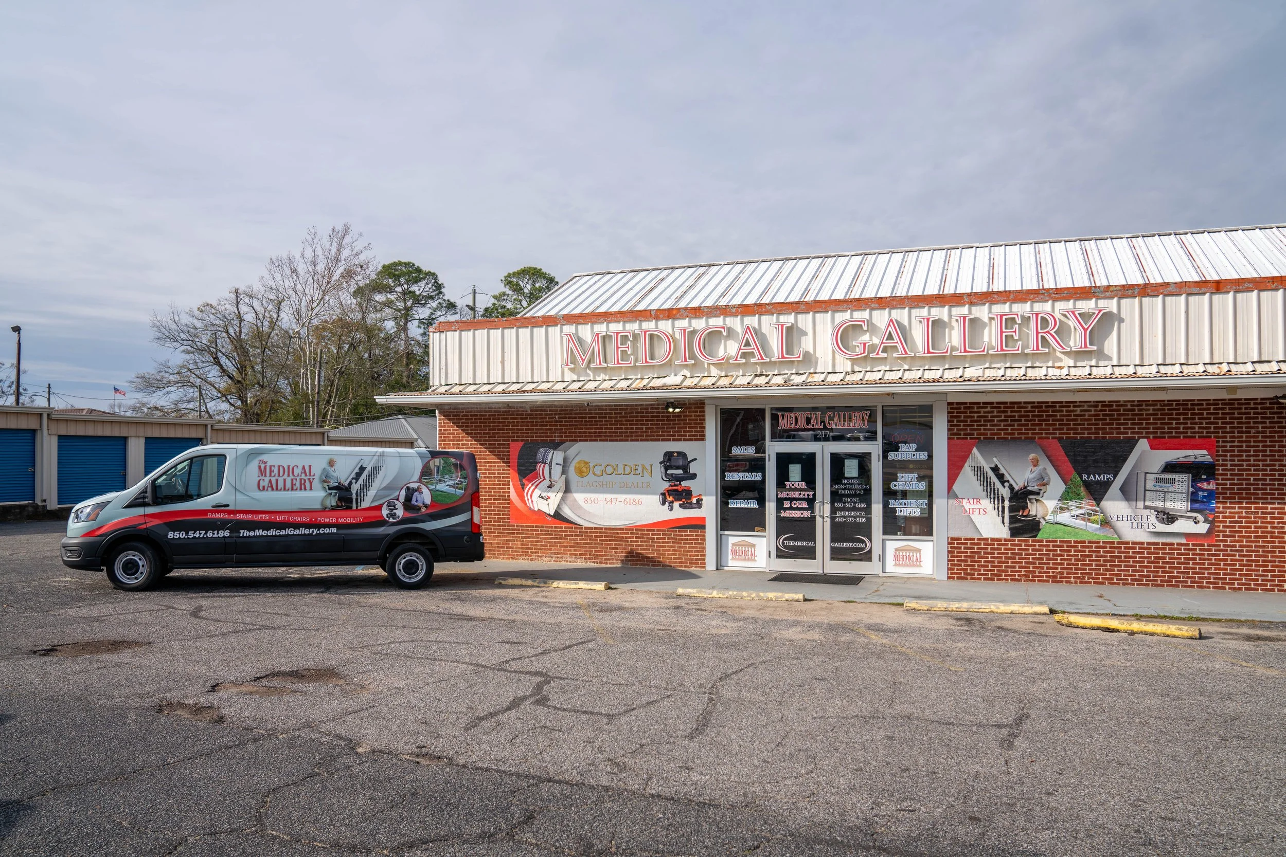 Exterior of a store called Medical Gallery with a delivery van parked outside. The store has a brick facade with promotional banners and signs on the windows displaying medical equipment like stair lifts, vehicle lifts, and other accessibility produc