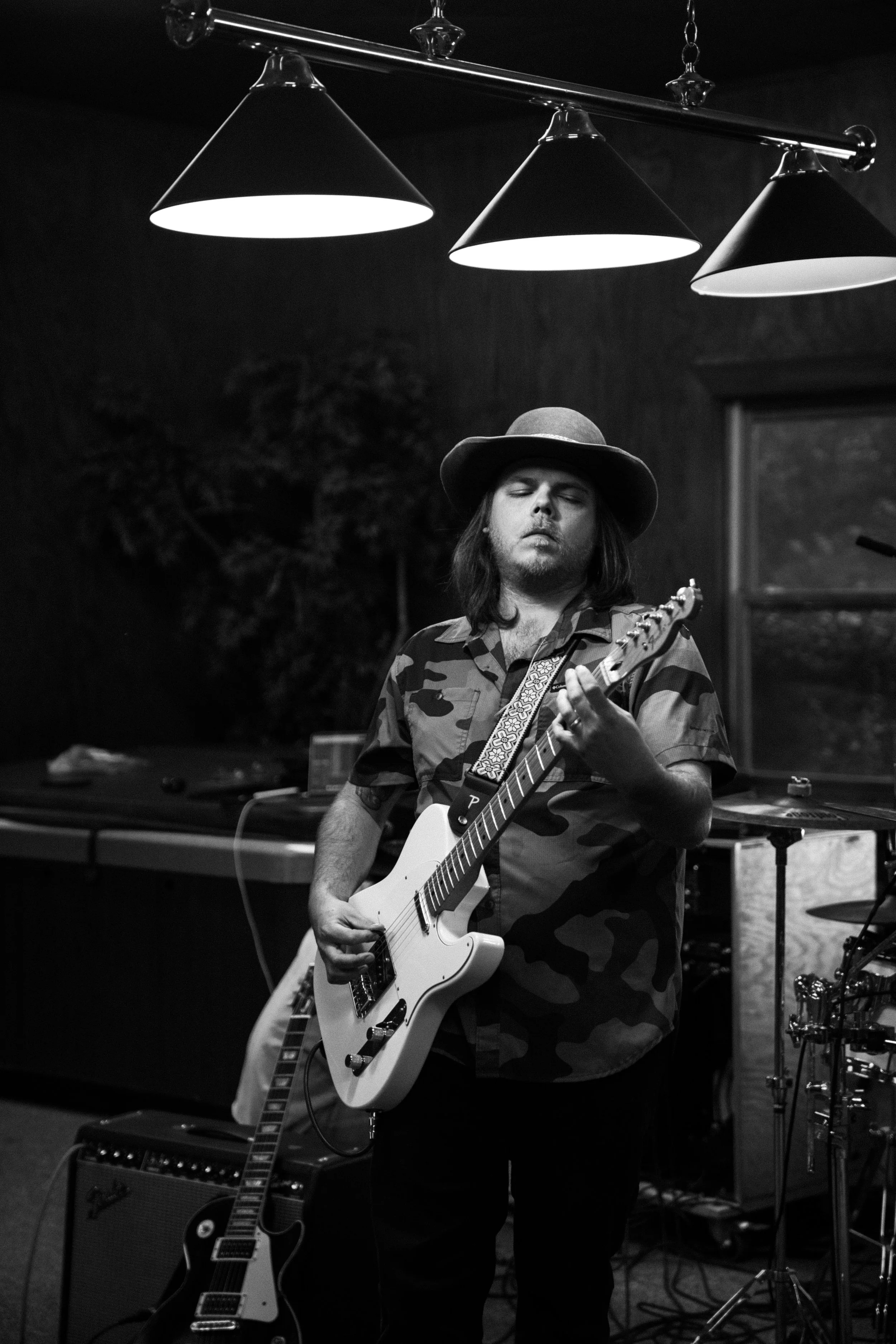 A man wearing a camo shirt and wide-brim hat playing an electric guitar in a dimly lit room.