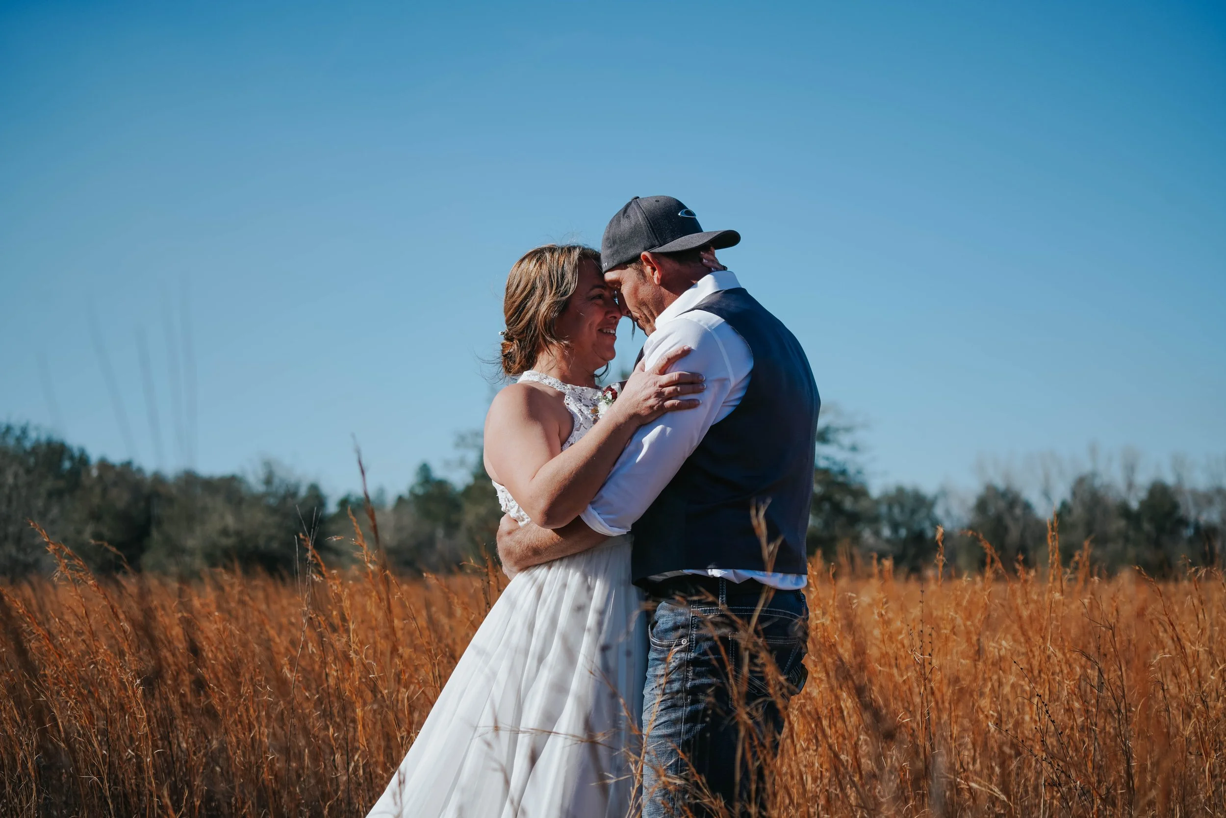 A woman in a white dress and a man in casual attire embracing and smiling in a field of tall grass under a clear blue sky.