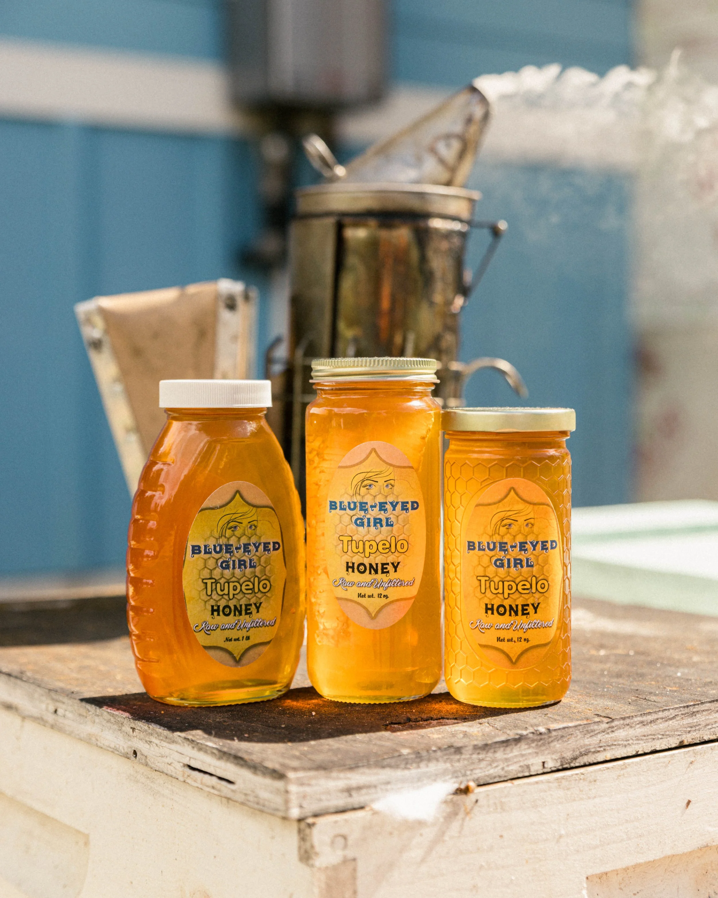 Three jars of Blue-Eyed Girl Tupelo Honey on a wooden surface, with a honey extractor in the blurred background.