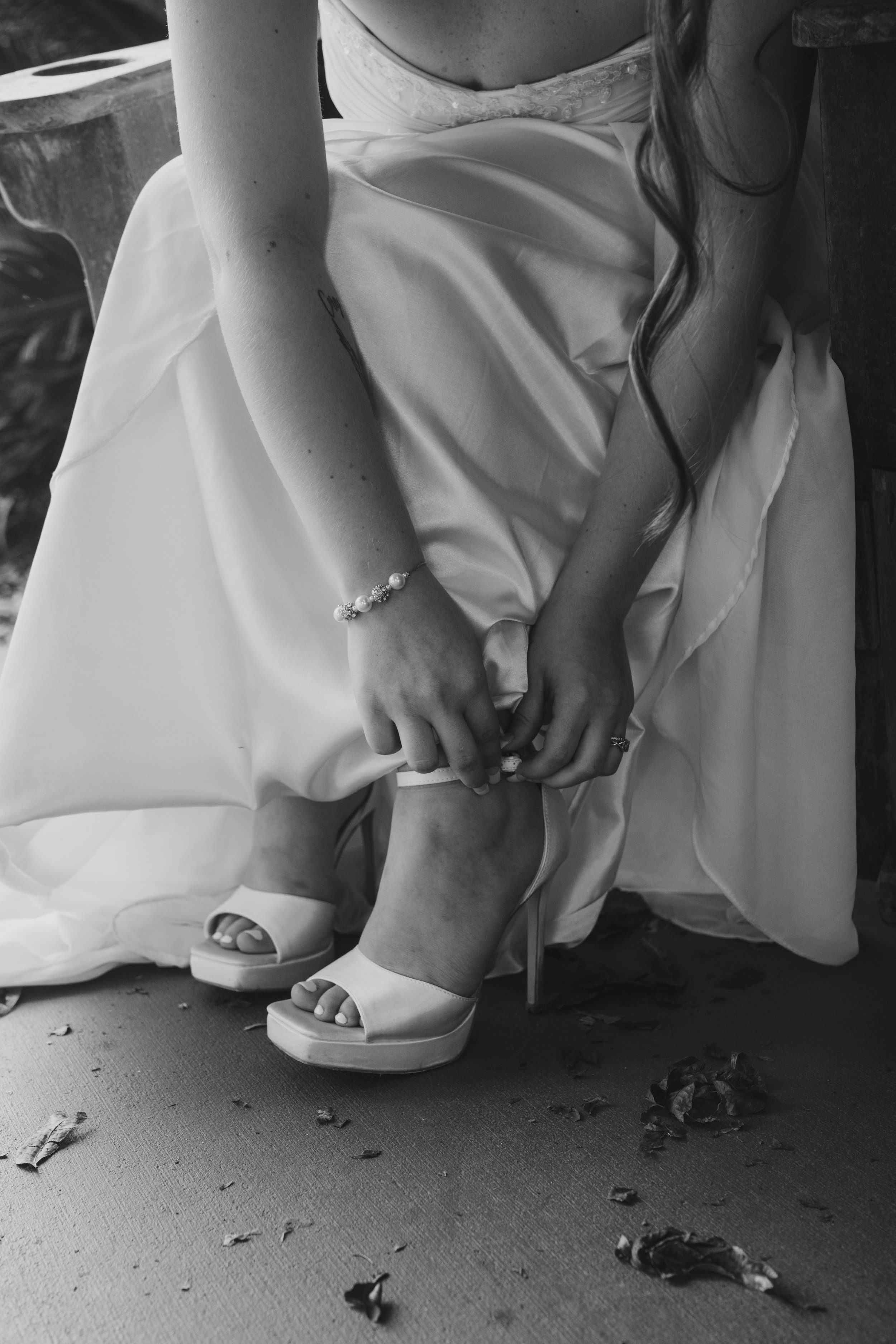 A bride sitting down, adjusting her high-heeled shoe, wearing a wedding dress and jewelry, with scattered rose petals on the floor.