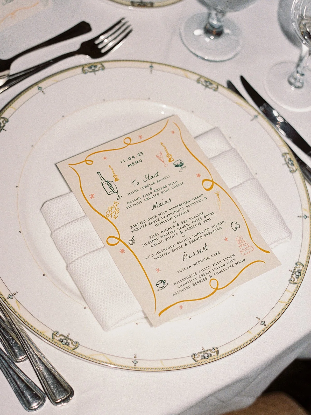 An illustrated, hand-drawn menu card nestled in a folded napkin atop monogrammed University Club china — playful stationery design meeting formal table setting in perfect balance.