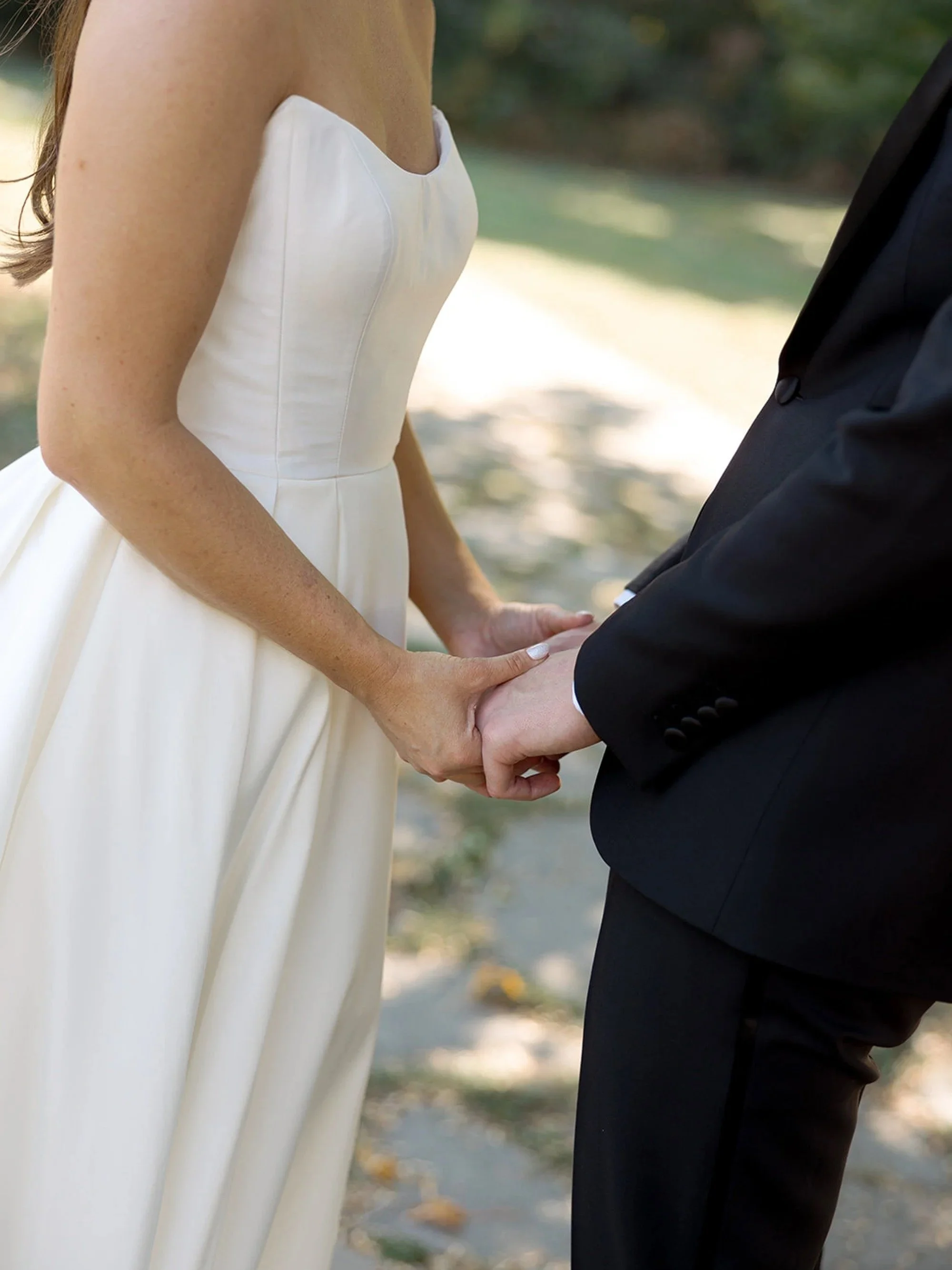 A quiet, intimate close-up — the bride and groom's hands intertwined on a sunlit garden path, their brand-new wedding rings visible. Columbus Park Refectory