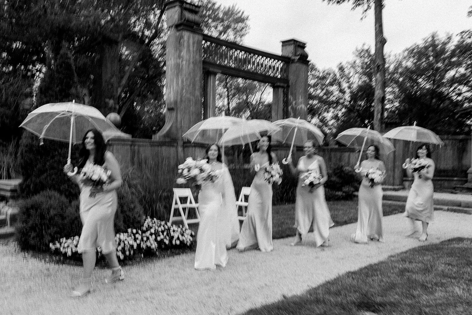 Black and white photo of bridesmaids in satin gowns walking through the rain with clear umbrellas at Armour House in Lake Forest, photographed by Chicago film wedding photographer Louie Abellera.