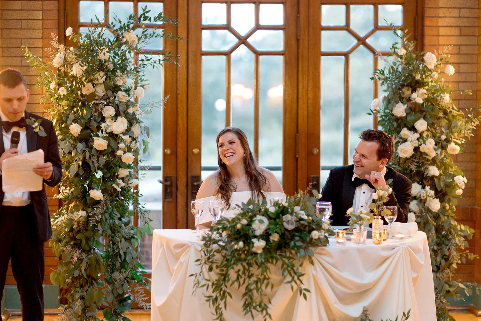 Bride bursting into laughter at the sweetheart table during a wedding toast, the groom grinning beside her.