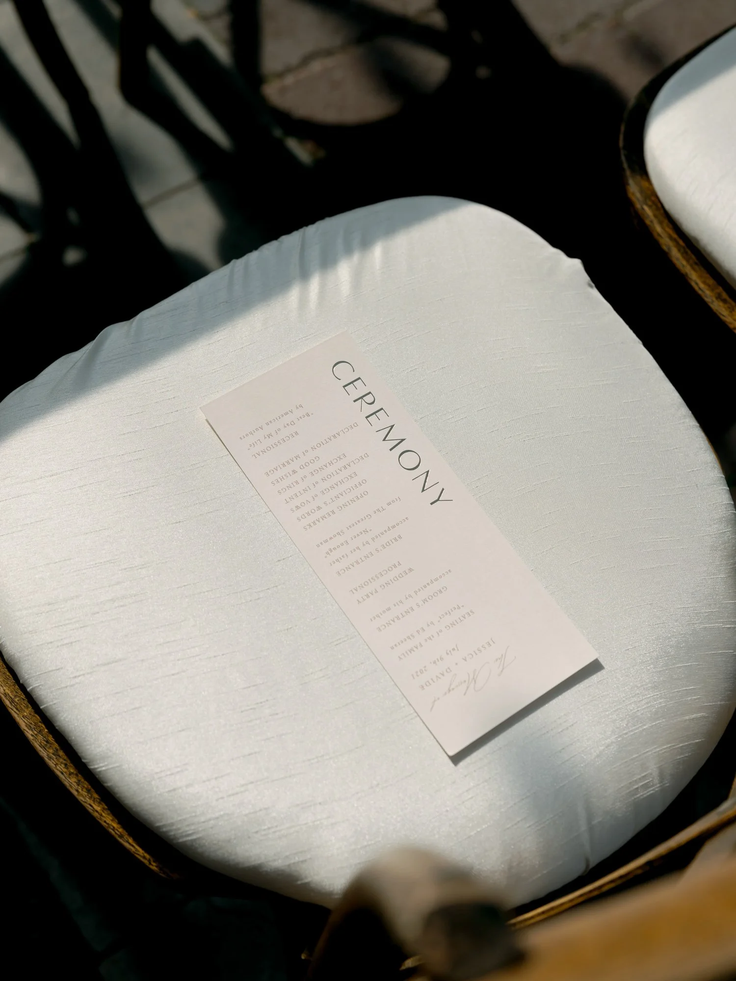 An elegant cream ceremony program rests on a white cushioned chair seat at the Chicago Illuminating Company, with dramatic geometric shadows cast by sunlight.