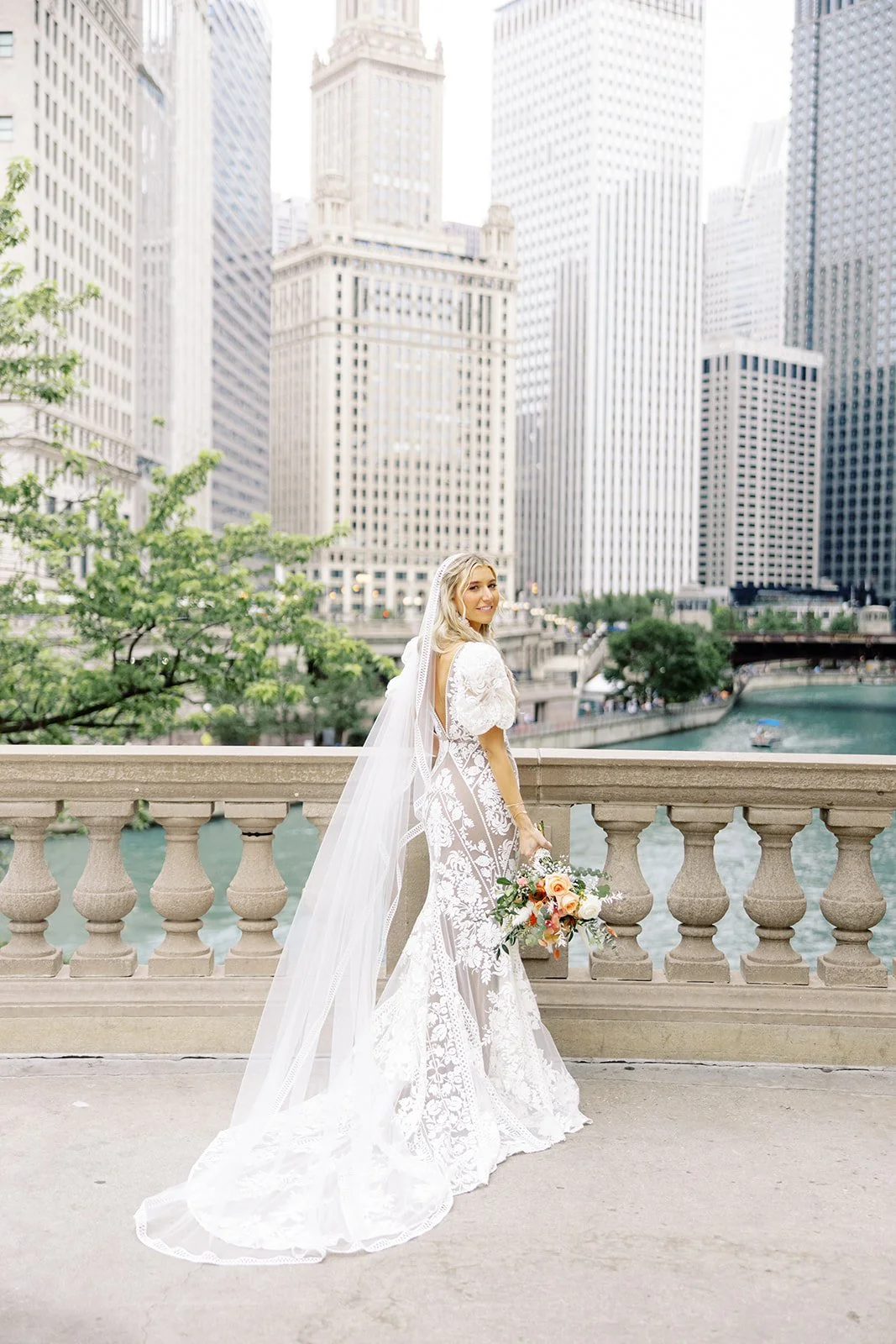 Bride in a flowing bohemian wedding dress standing in front of the Wrigley Building on the Chicago Riverwalk, captured by Chicago wedding photographer Louie for a Room 1520 wedding.