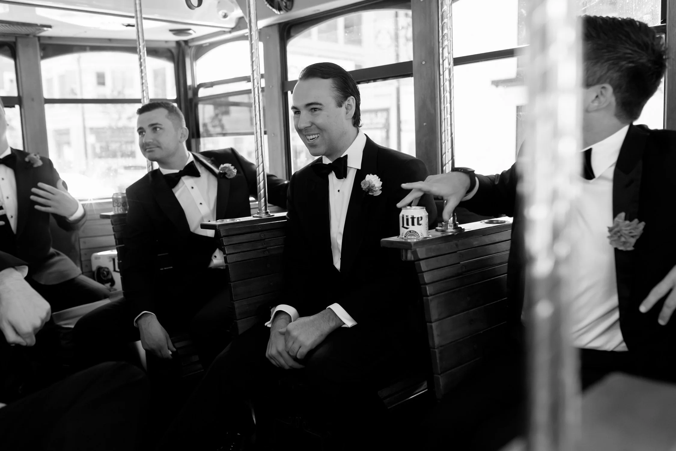 Groom and groomsmen in black tuxedos ride a vintage Chicago trolley in black and white, laughing with a can of Miller Lite on the armrest between them.
