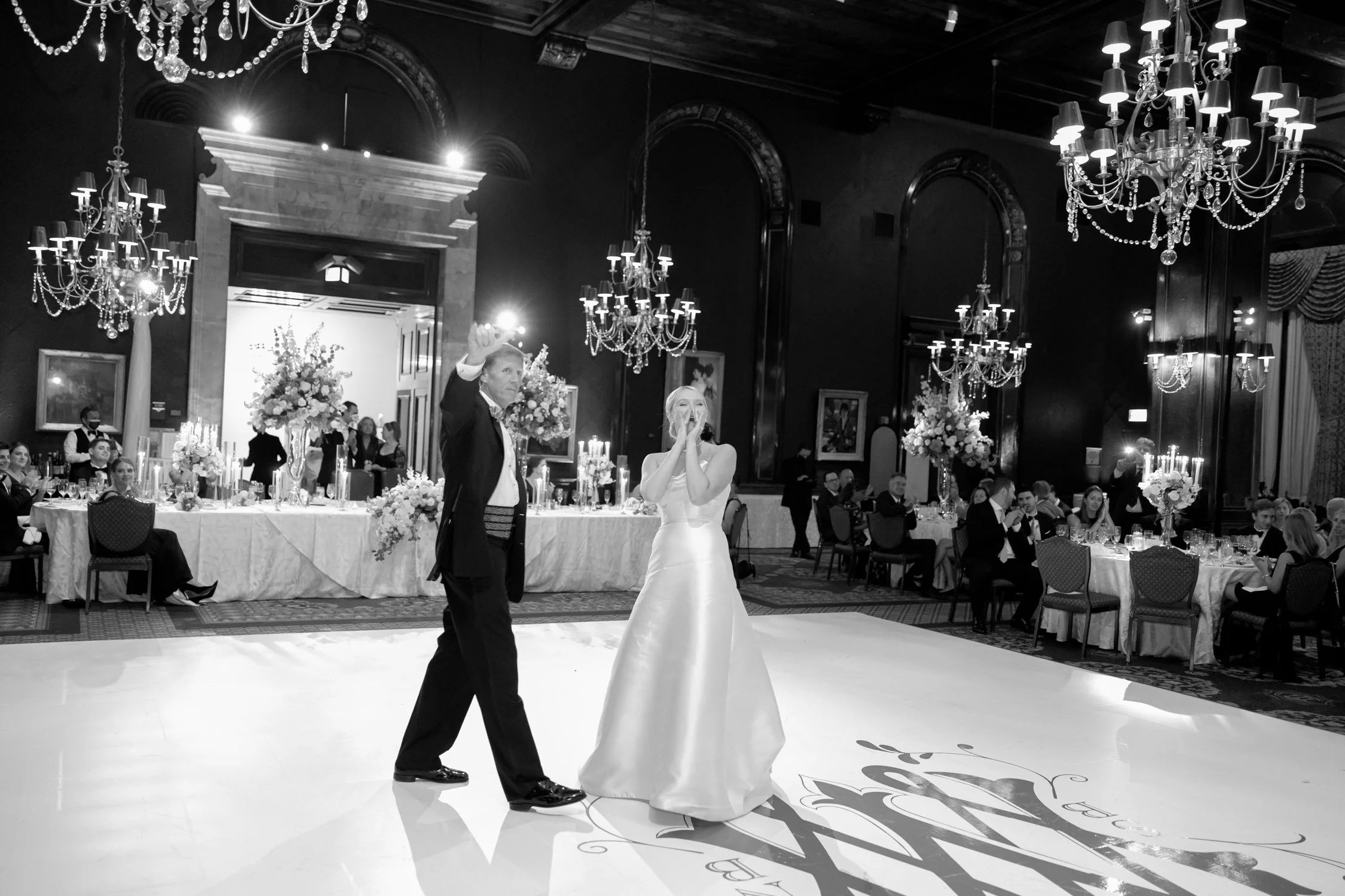 Wide black and white shot of the father-daughter dance in the Union League Club grand ballroom — a custom gold monogram floor lit beneath their feet.
