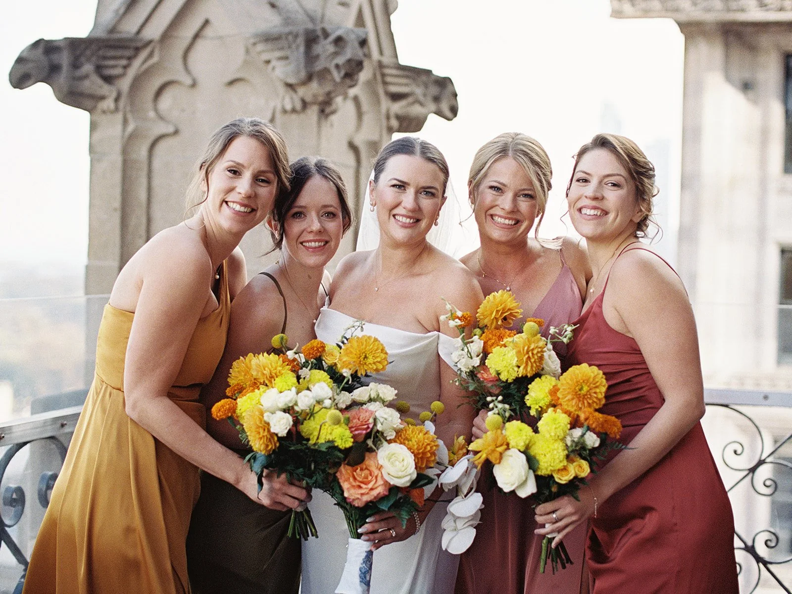 The bride and her four bridesmaids on the University Club Chicago rooftop terrace, all smiling and laughing, Gothic stone spires rising dramatically behind them against an open sky.