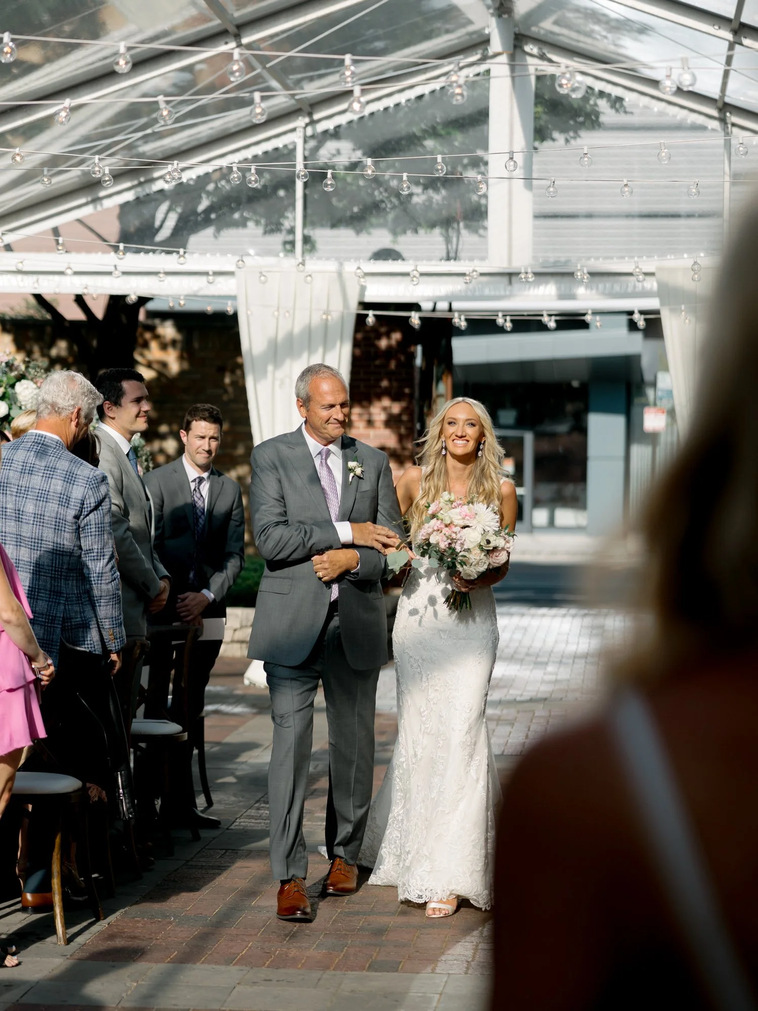 The bride radiates joy as her father escorts her down the aisle of the Chicago Illuminating Company ceremony, guests rising and smiling all around them.