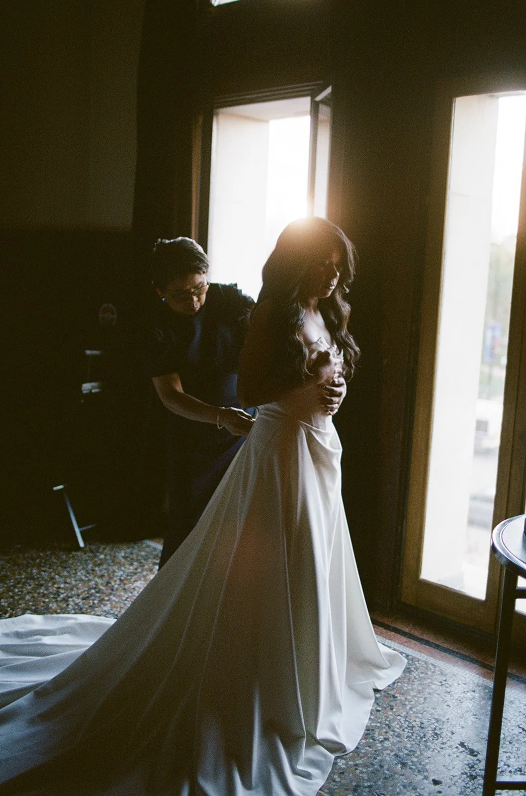 Bride Sarah being laced into her white gown in a darkly lit room at Chicago Athletic Association, backlit by elegant windows. Shot on film by a Chicago film wedding photographer.