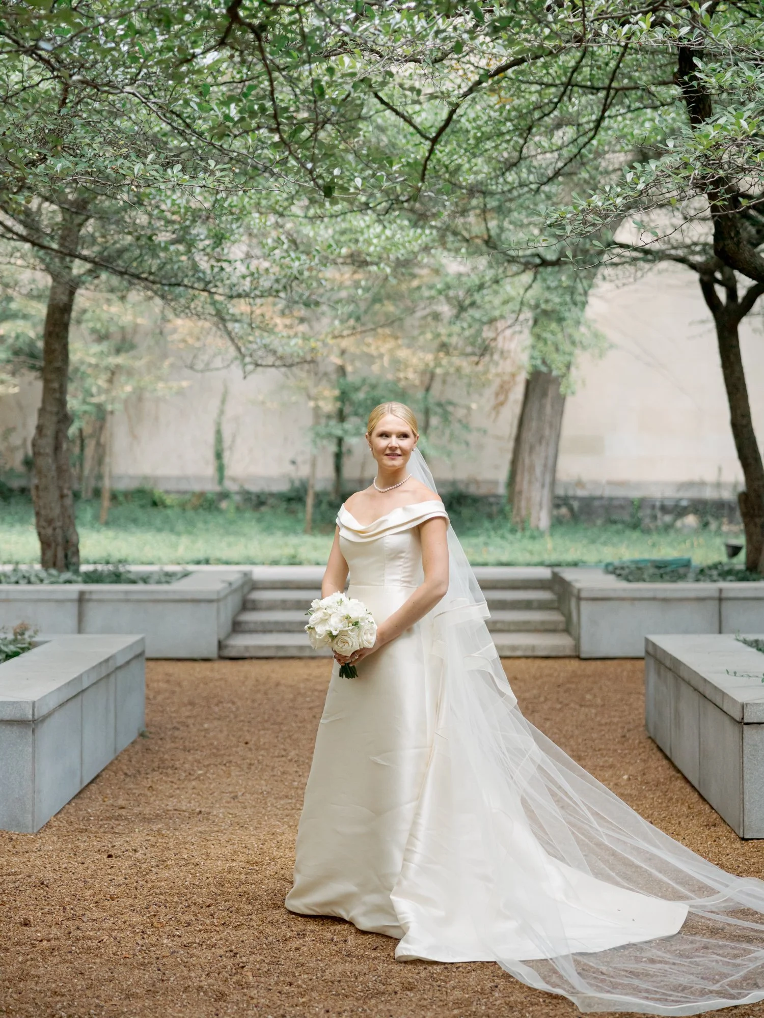 Full-length bridal portrait in the tree-lined garden courtyard — off-shoulder silk gown, cathedral veil trailing, and a clean white bouquet in hand.
