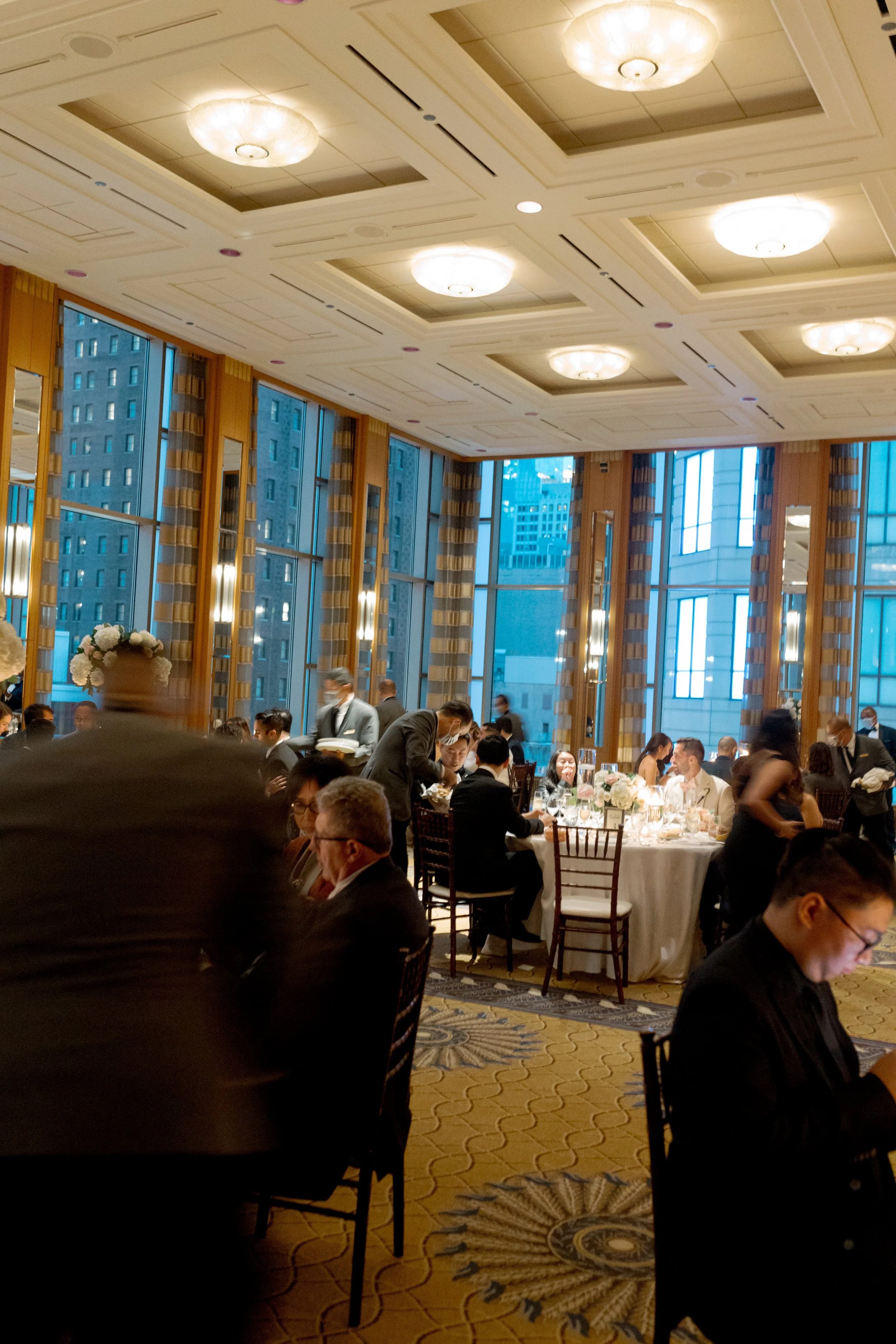 Wide view of the Peninsula Chicago reception hall with guests dining, florals and floor to ceiling city skyline windows