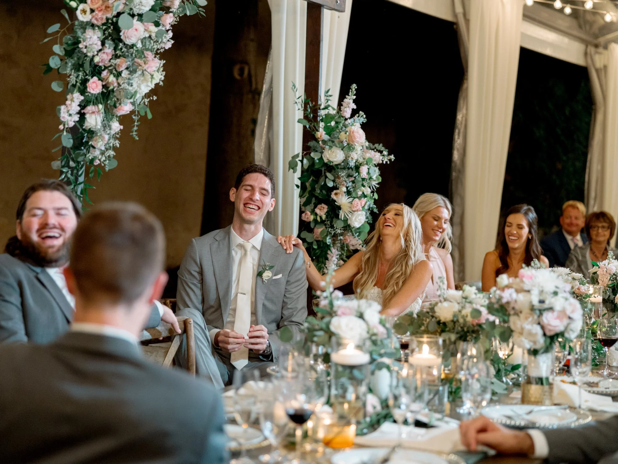 The bride and groom erupt in laughter at the sweetheart table during speeches at the Chicago Illuminating Company, surrounded by greenery and candlelight.