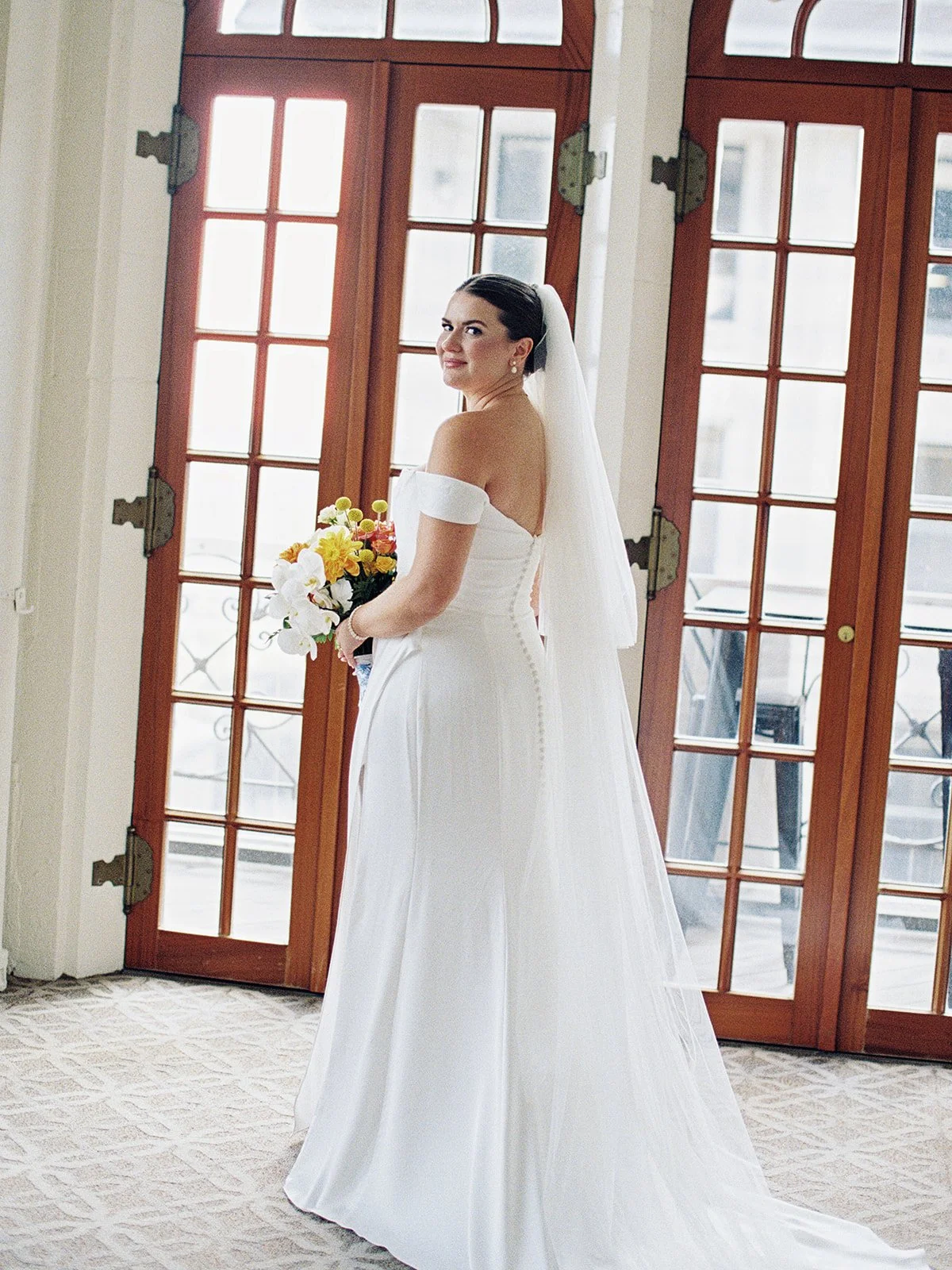 The bride turns to look over her shoulder inside the ornate University Club, bouquet in hand, French doors flooding the room with soft city light behind her. A bridal portrait that feels effortless and editorial