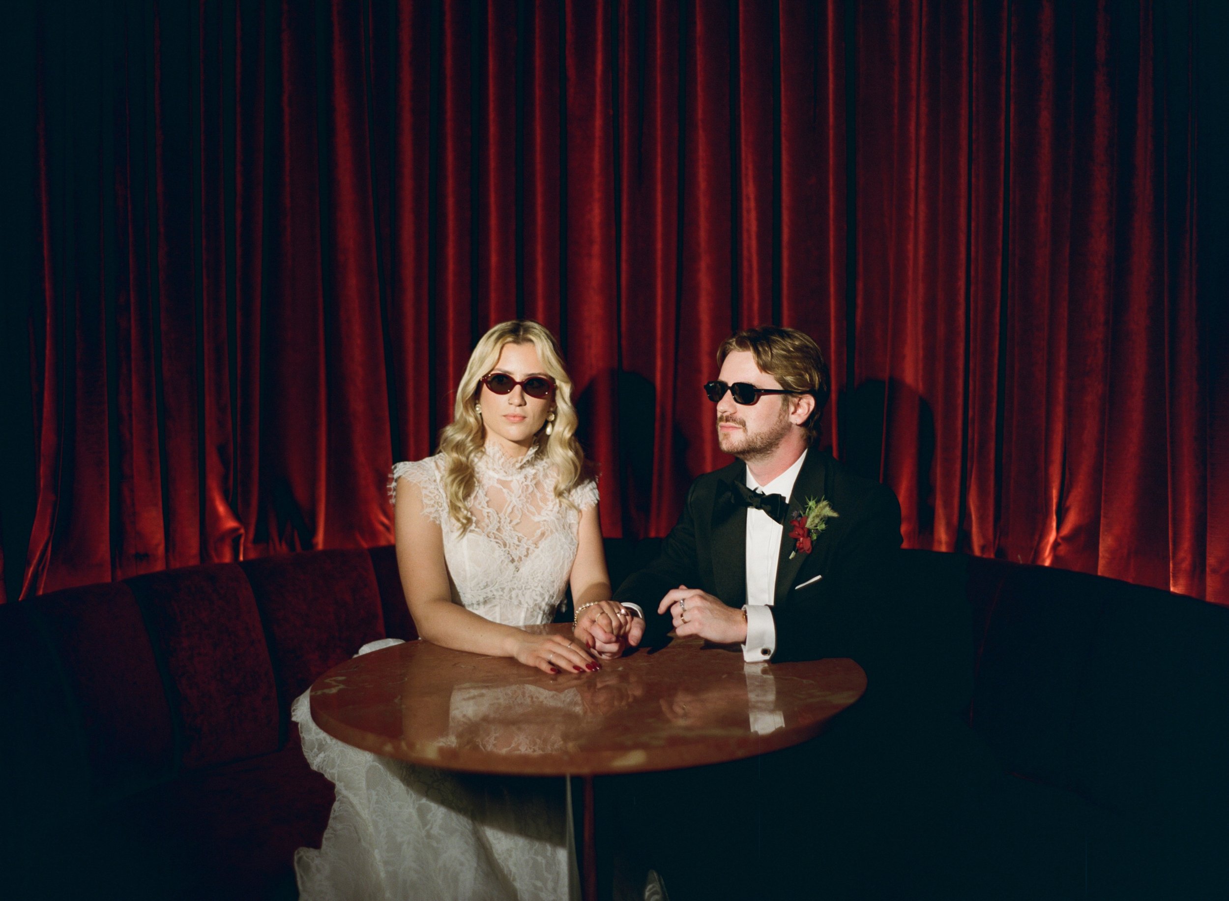 Bride and groom in sunglasses hold hands at a round table inside The Wellsley's red velvet lounge