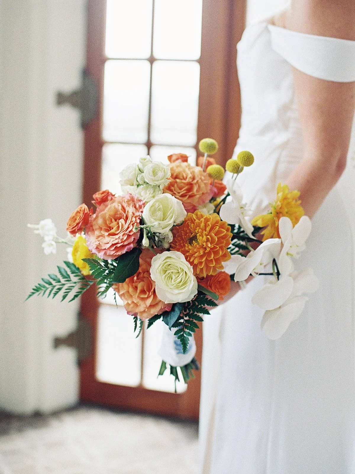 The bridal bouquet in close-up: coral ranunculus, orange dahlias, white orchids, and yellow billy balls held against the crisp off-shoulder gown — a burst of saturated color against clean white. Shot on film by a Chicago film wedding photographer.