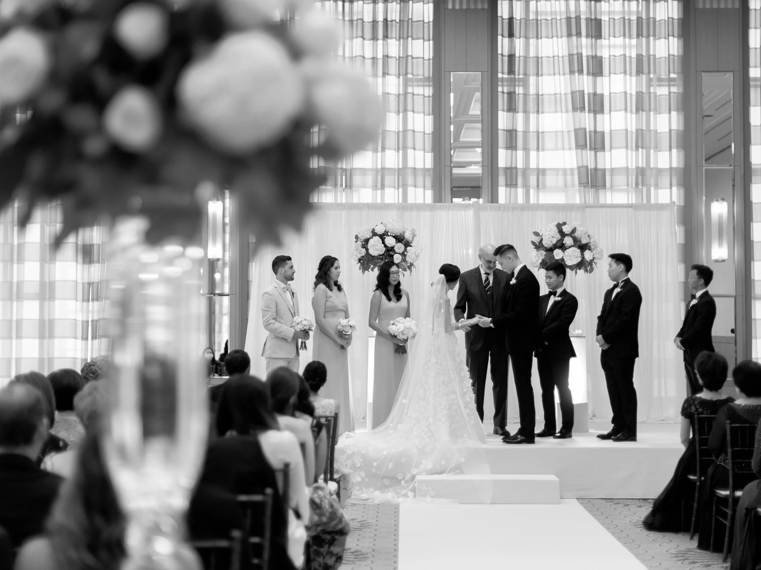 Black and white wide photograph of the wedding ceremony framed by florals and skyline windows at The Peninsula Chicago